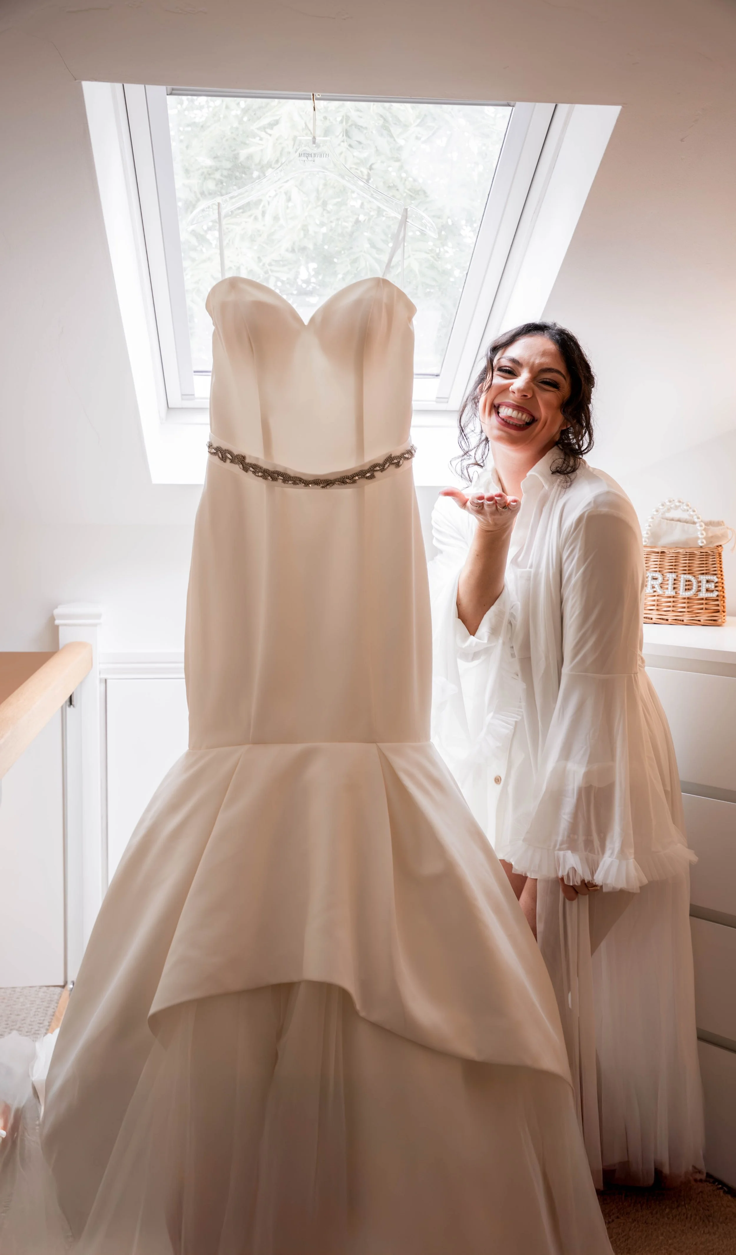 A smiling woman with dark curly hair in a white shirt poses next to a hanging wedding dress in a room with a skylight window.