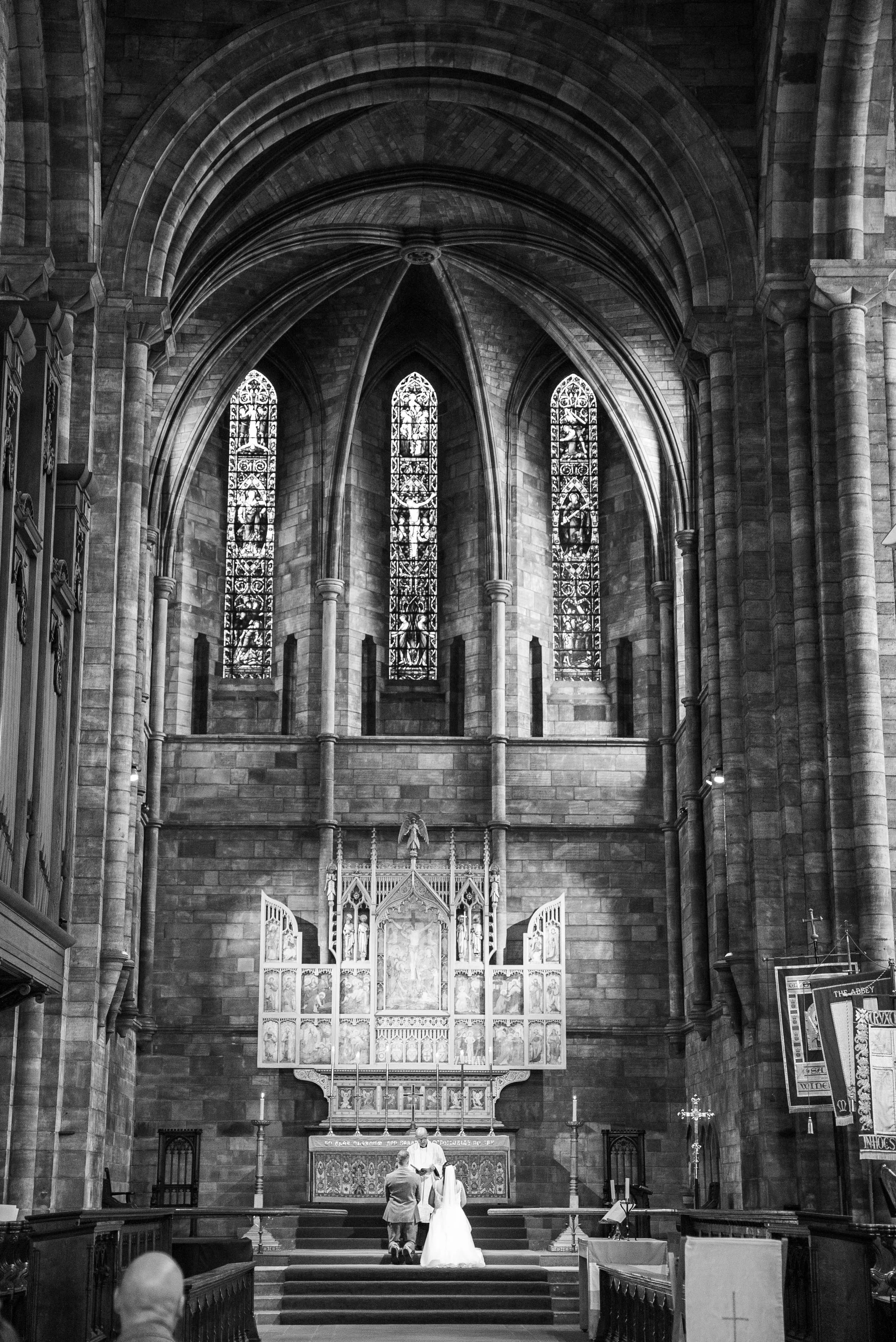 A black and white photo of a church interior showing the altar with a couple getting married, clergy officiating, and stained glass windows.