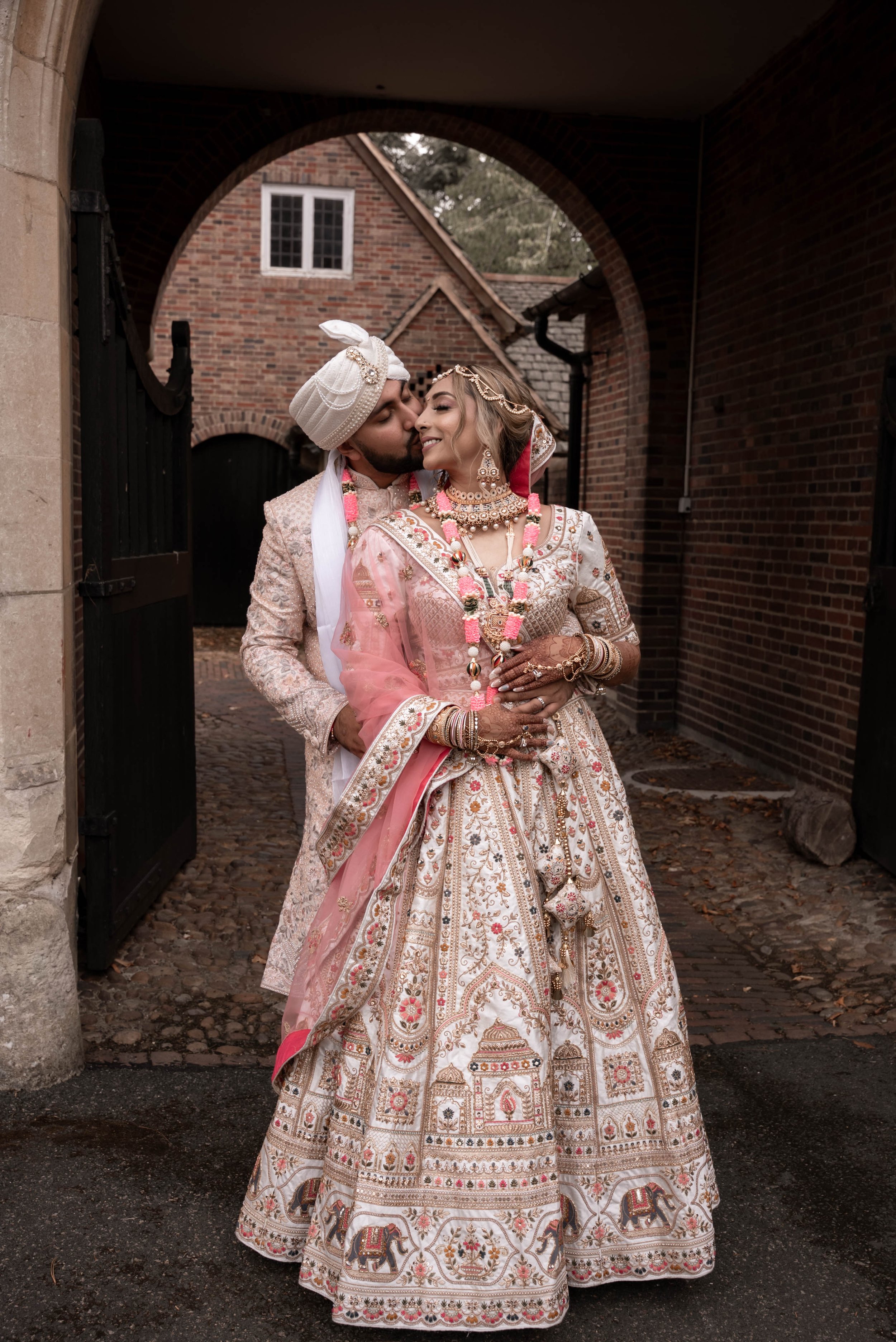 Bride and groom dressed in traditional Indian wedding attire sharing a romantic moment under an archway in a brick courtyard.