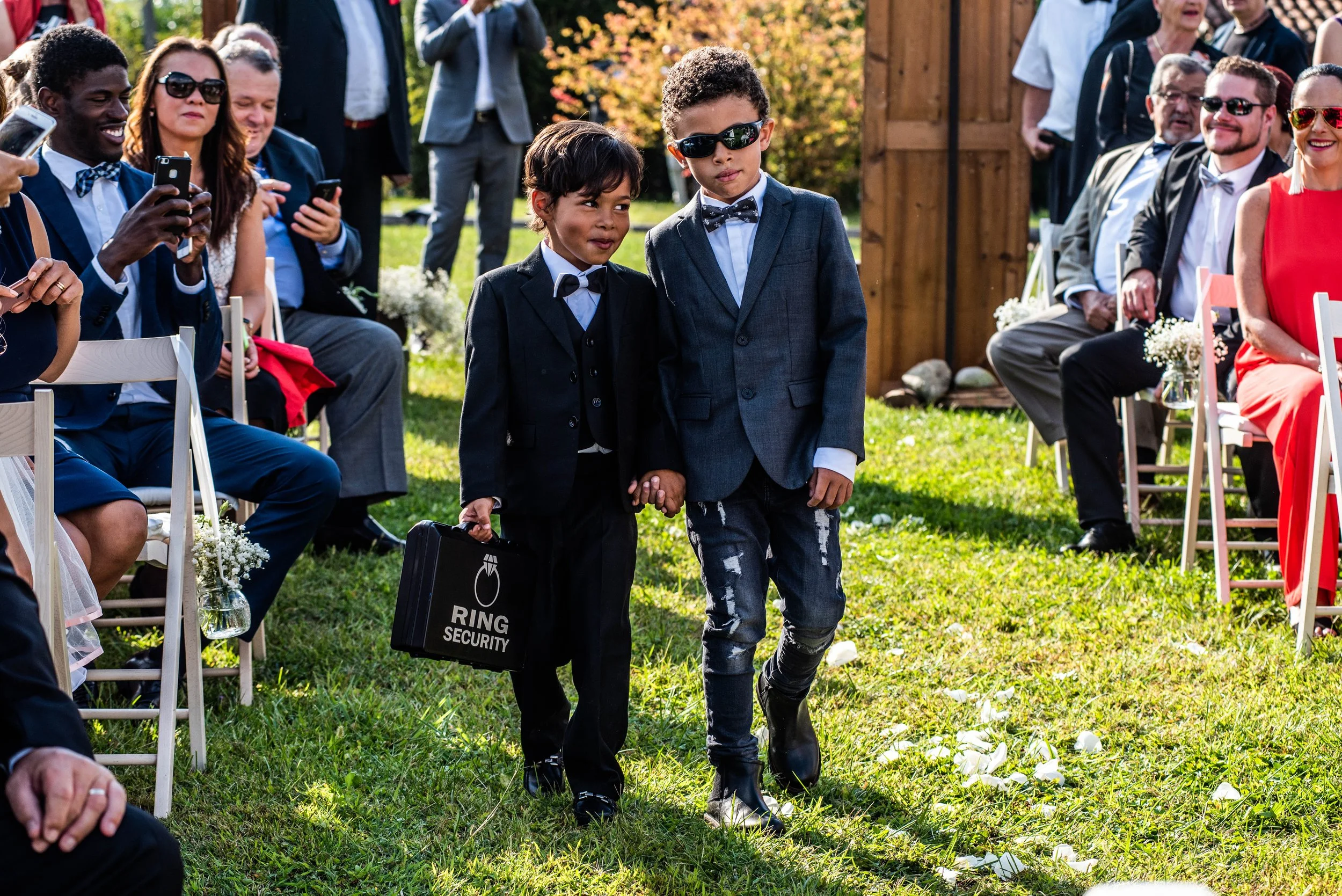 Two young boys dressed in suits walking hand-in-hand at an outdoor wedding ceremony, surrounded by seated guests taking photos and watching.