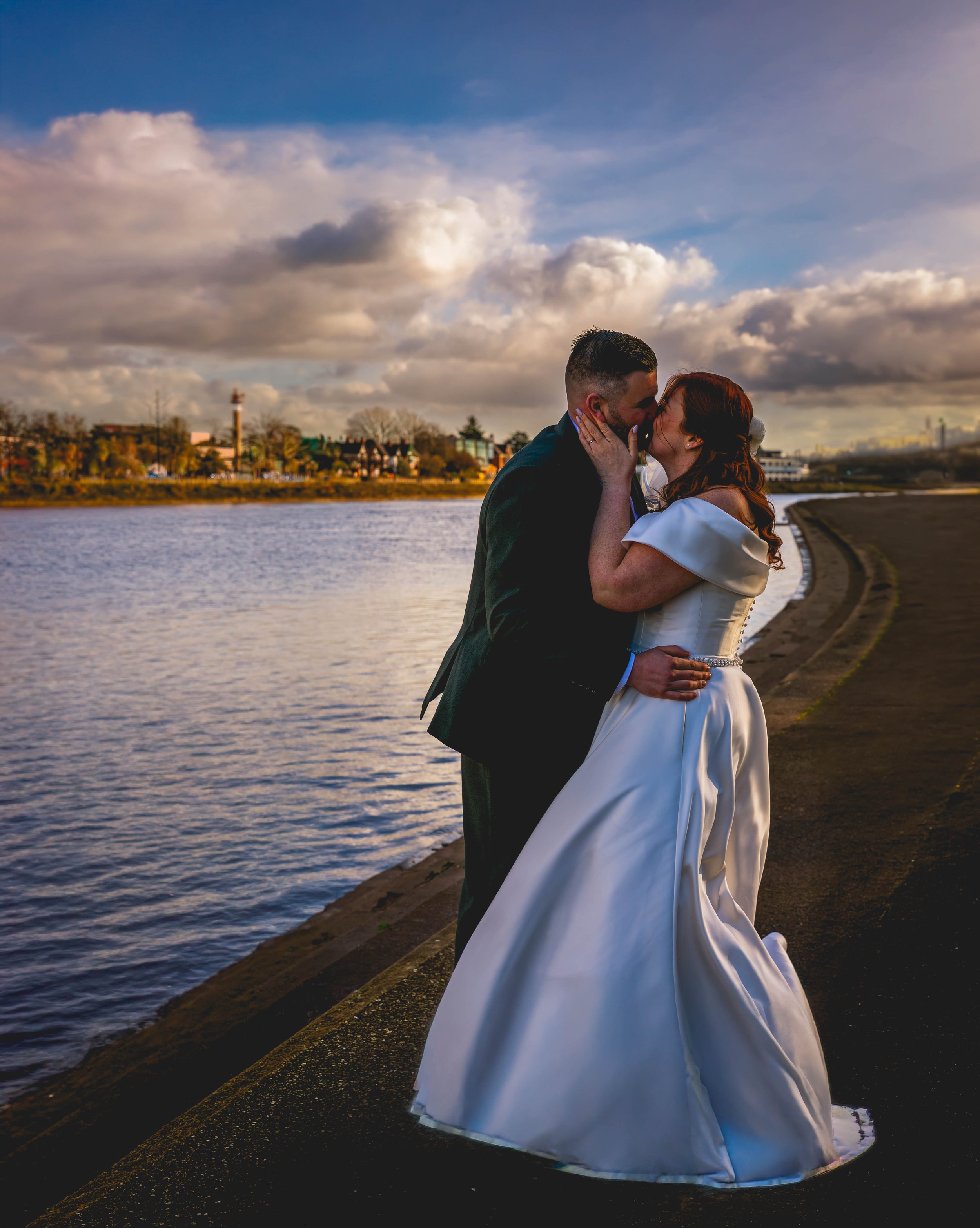 A newlywed couple kissing by a river during sunset, with clouds in the sky and city skyline in the background.