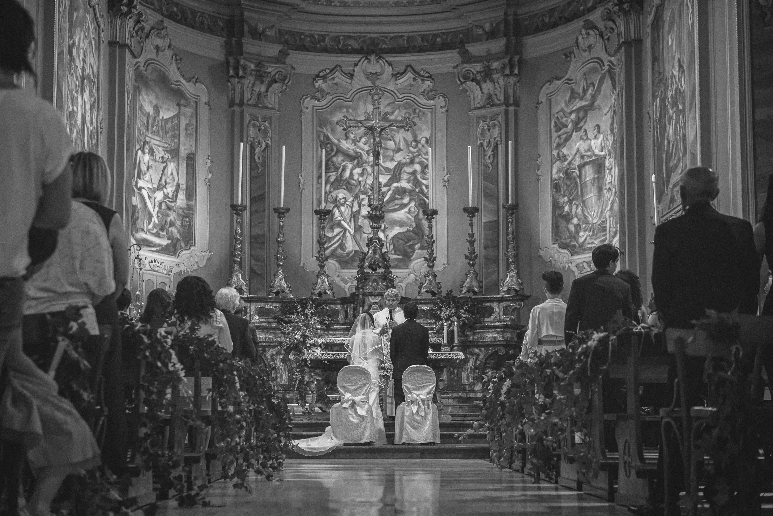 A black and white photo of a church or cathedral wedding ceremony. The altar features religious artwork and a crucifix above. The bride and groom are kneeling at the altar, facing a priest or officiant. Guests are seated and standing on either side, watching the ceremony.