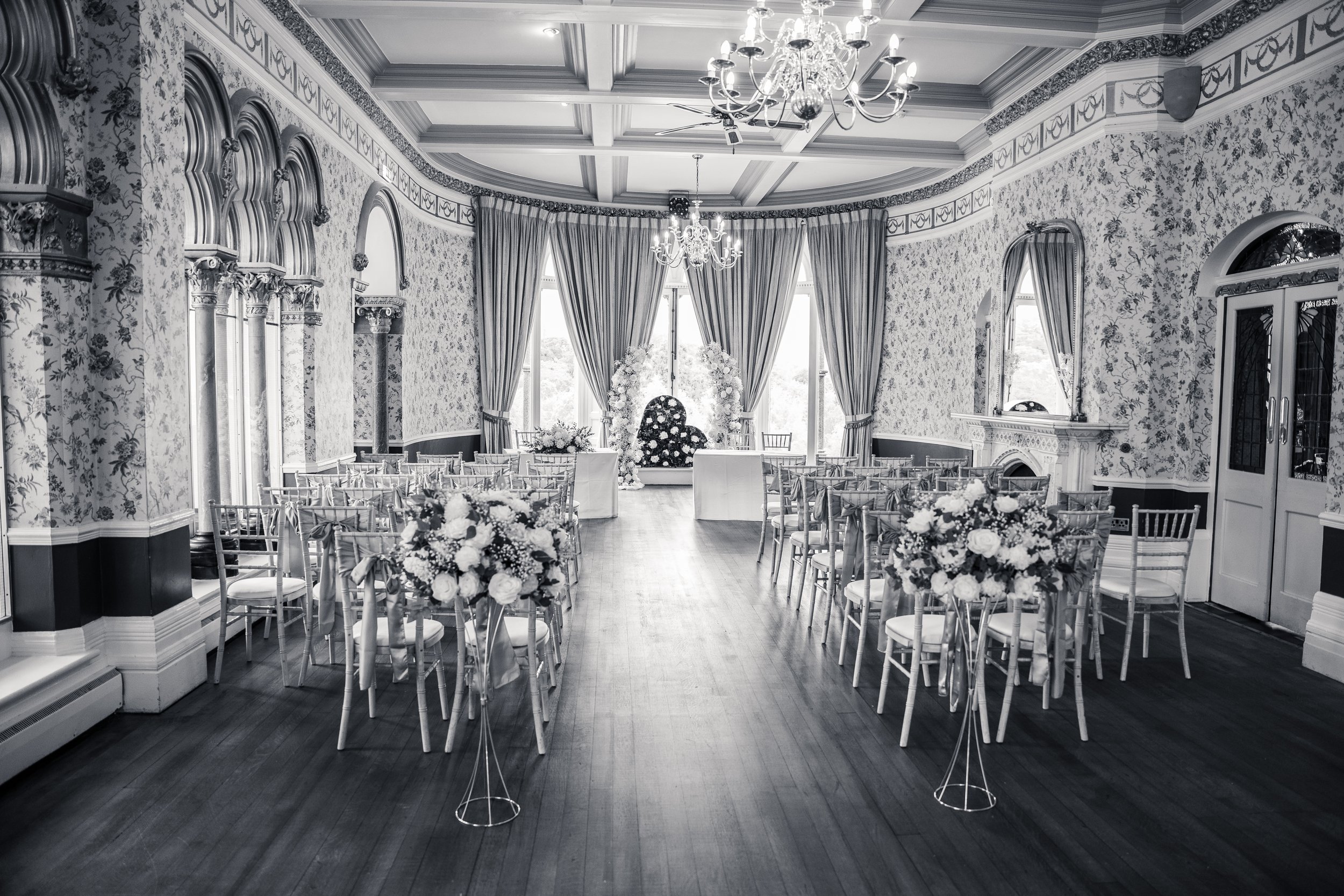 An elegant indoor event space decorated for a wedding or special occasion with floral arrangements, draped curtains, chandeliers, and rows of chairs.