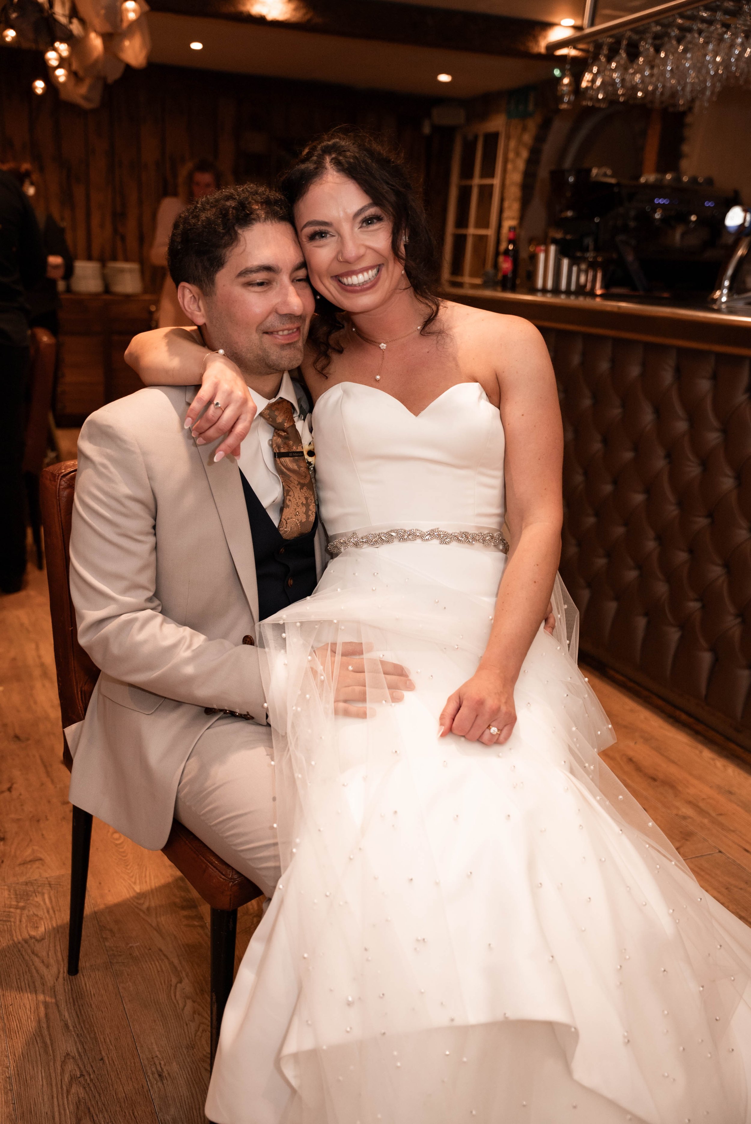 A bride and groom sitting together at their wedding reception, smiling and embracing. The bride is wearing a strapless white wedding gown with a beaded belt, and the groom is in a light beige suit with a patterned tie. The background shows a dimly li