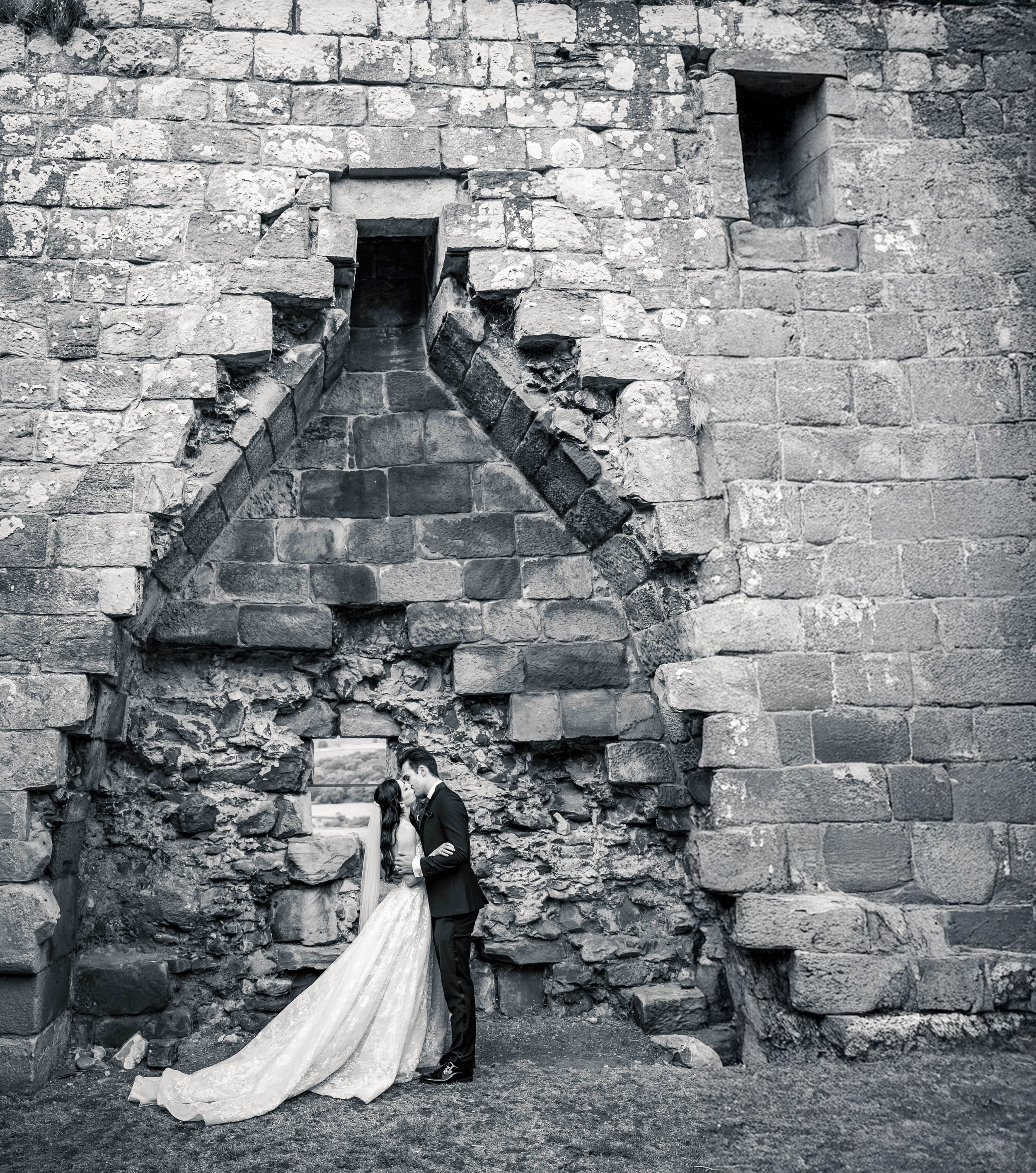 A black and white photo of a couple in wedding attire standing close together, embracing in front of an ancient stone wall with small windows and a large arched opening.