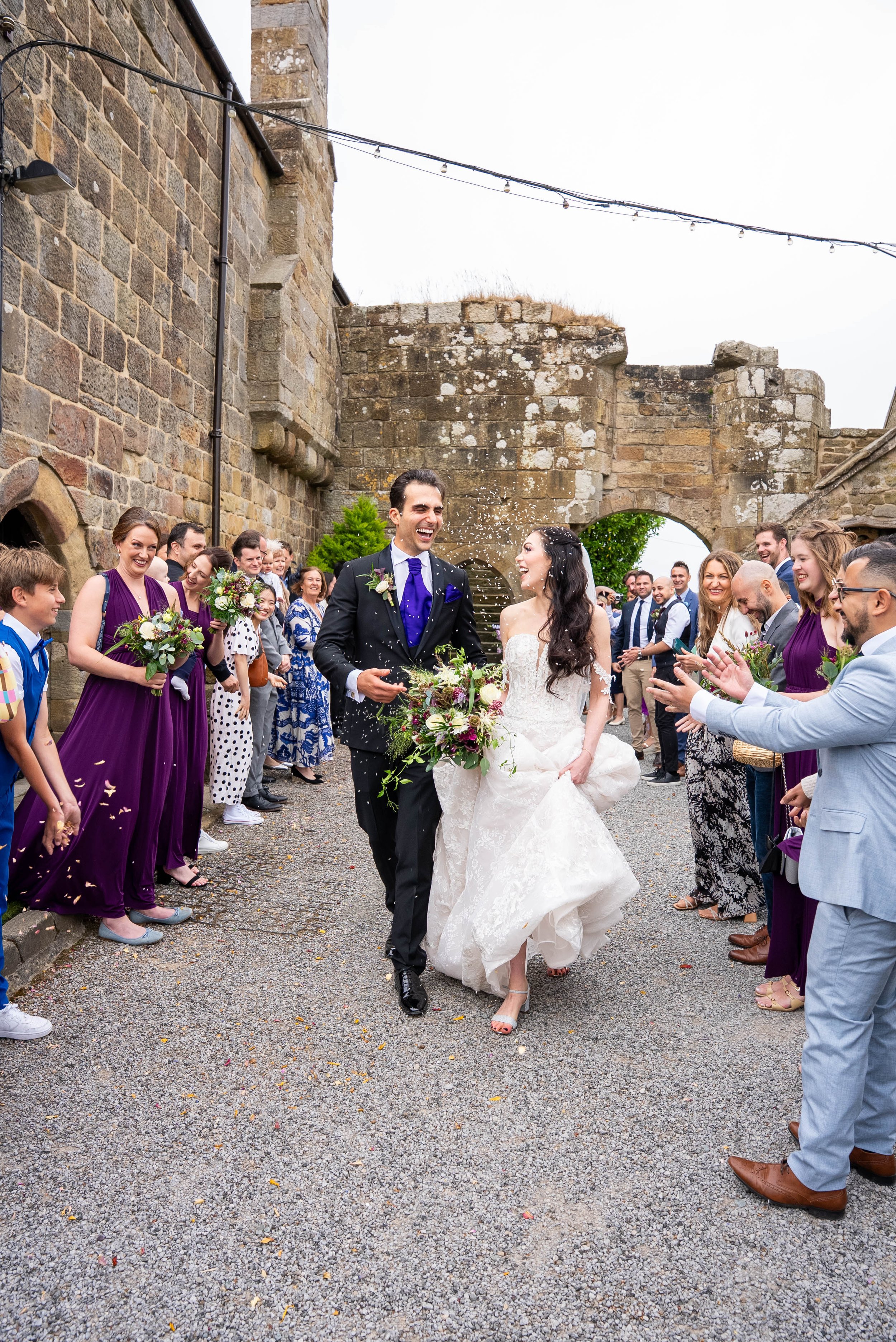 A newlywed couple walking together outside, surrounded by friends and family celebrating their wedding, with confetti falling from above.