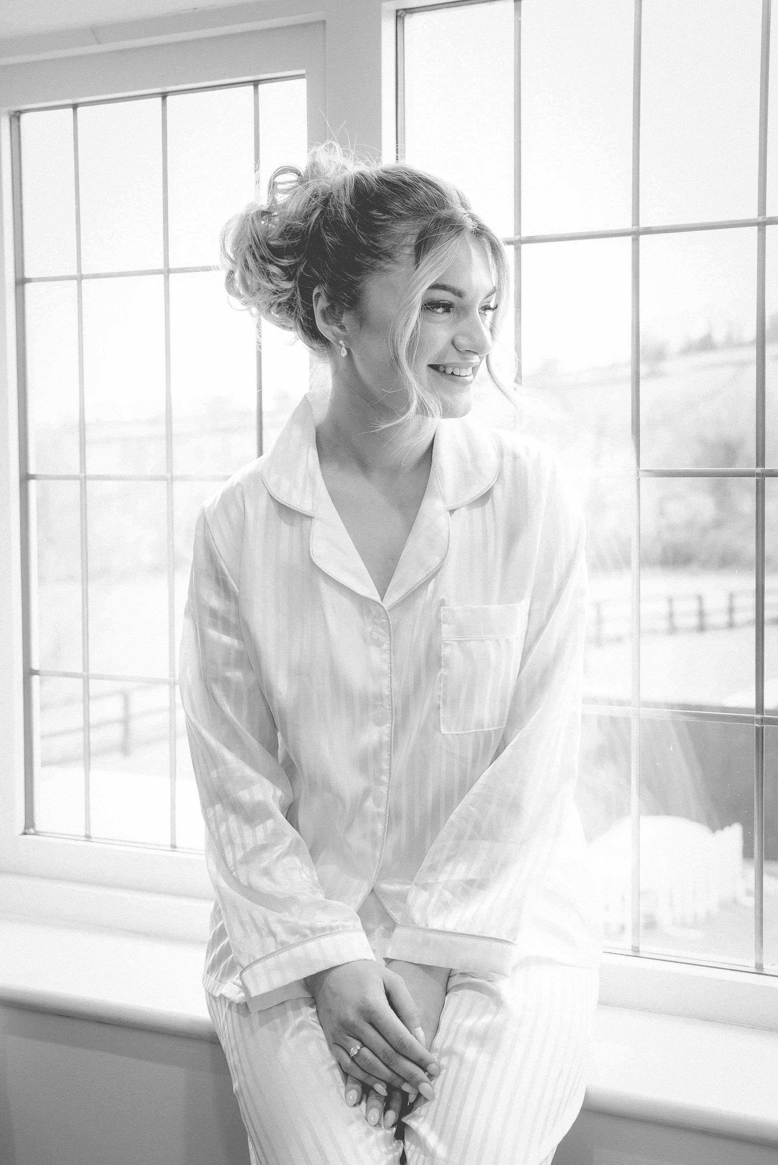 A woman in pajamas sitting by a large window, smiling, with her hands resting on her lap.