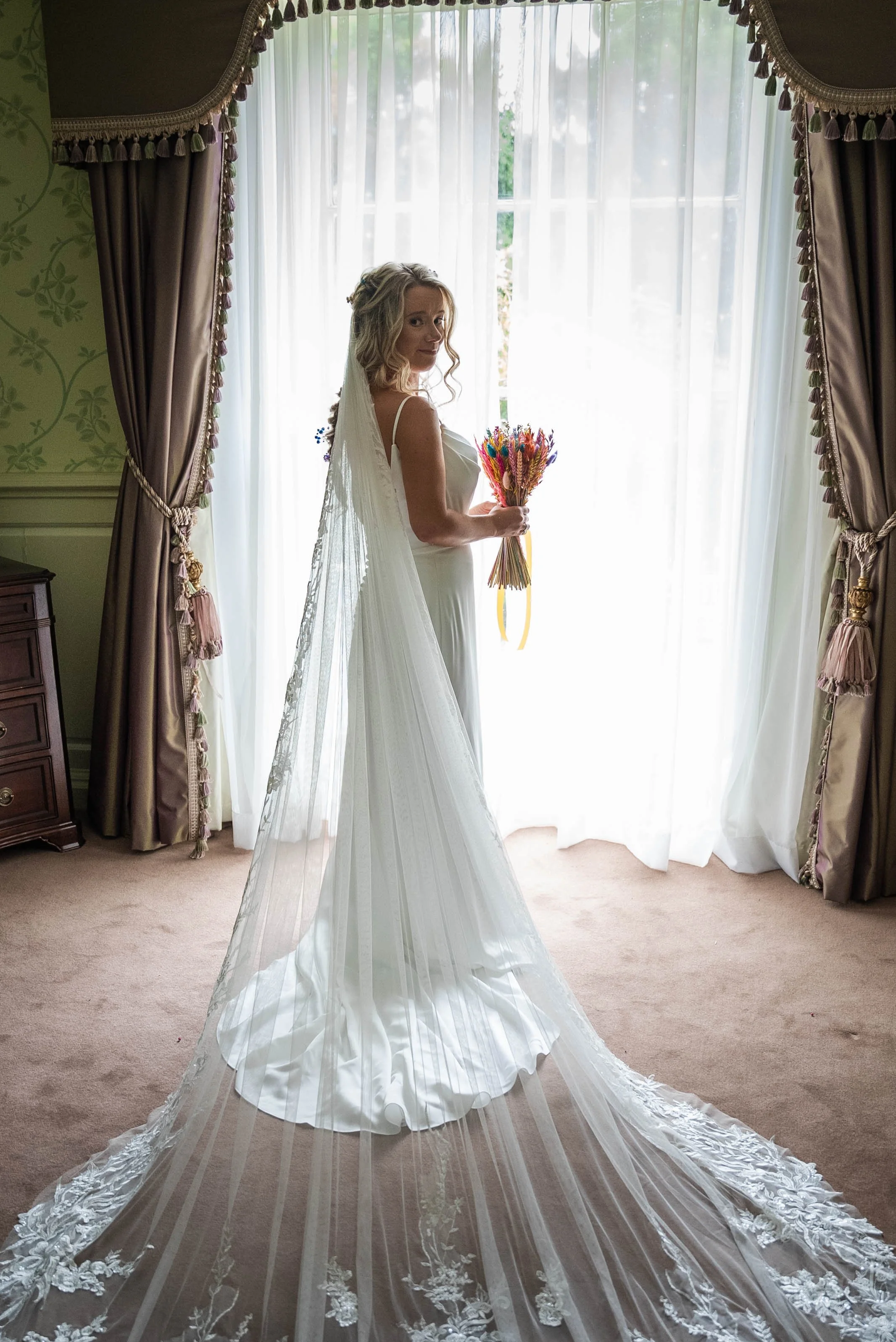 A bride in a white wedding dress with a long train and veil holding a bouquet of colorful dried flowers, standing in front of a window with sheer curtains and ornate curtains on either side.