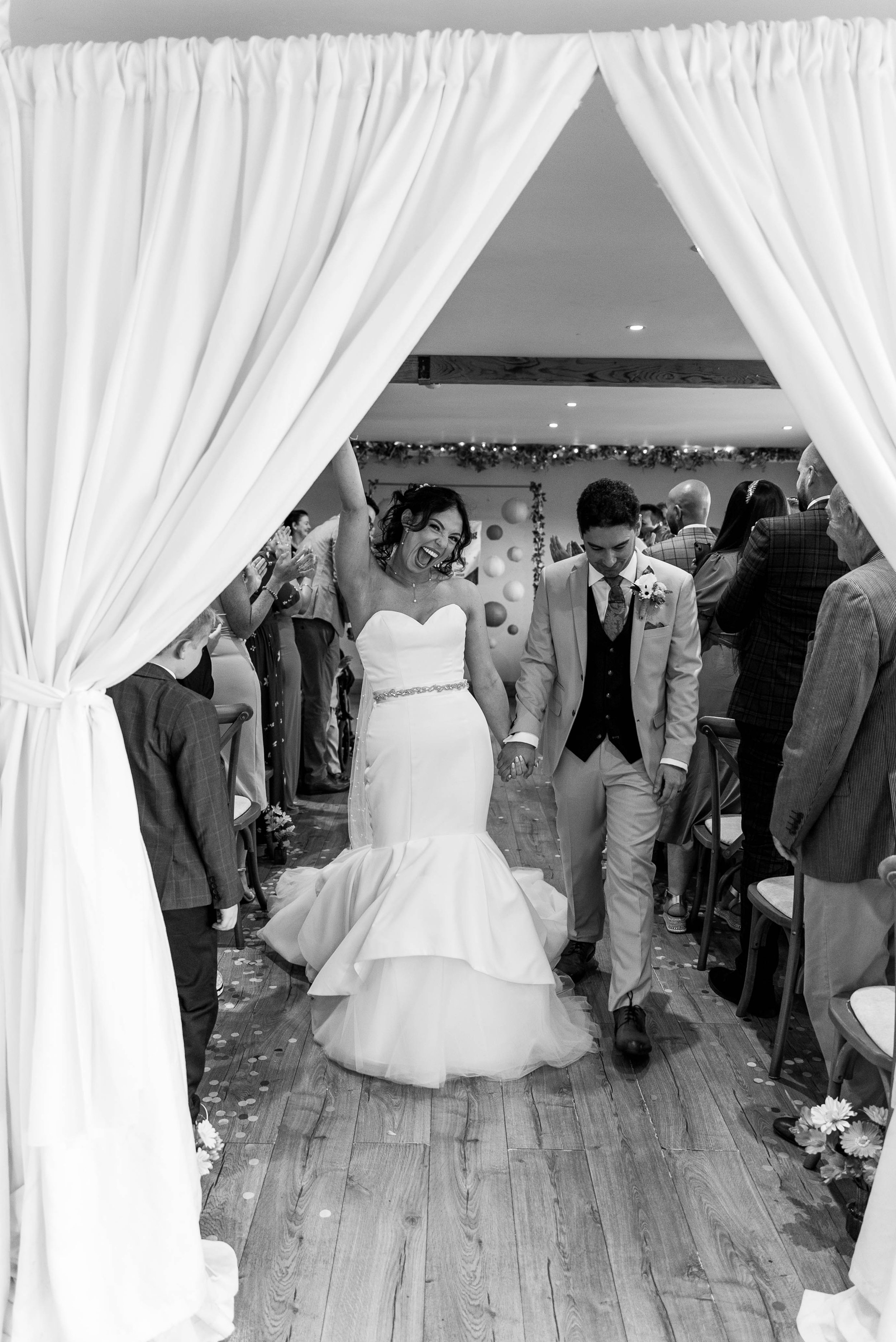 Bride and groom walking hand in hand through a wedding aisle, smiling, with wedding guests in the background.