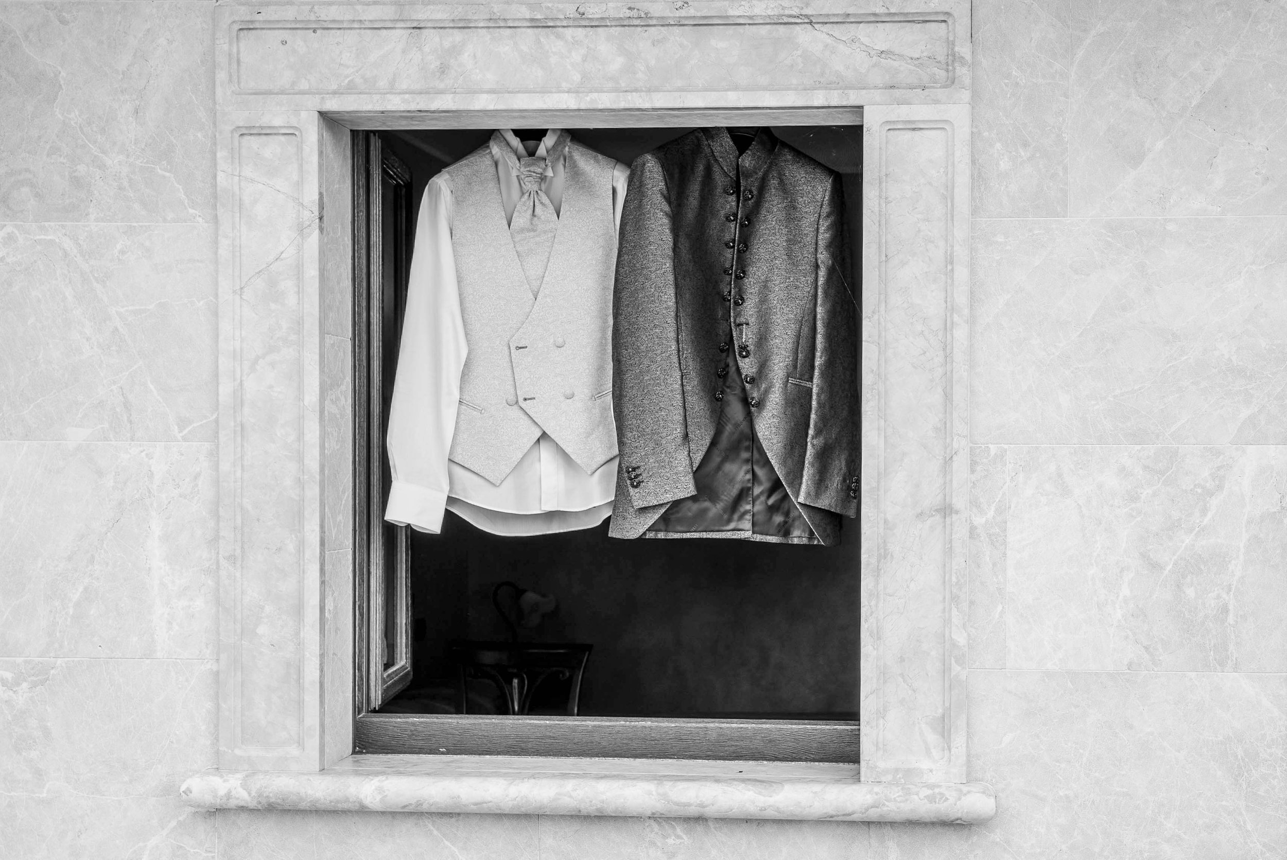 Black and white photograph of two suits hanging inside an elevator, seen from below. The elevator is surrounded by a marble wall.