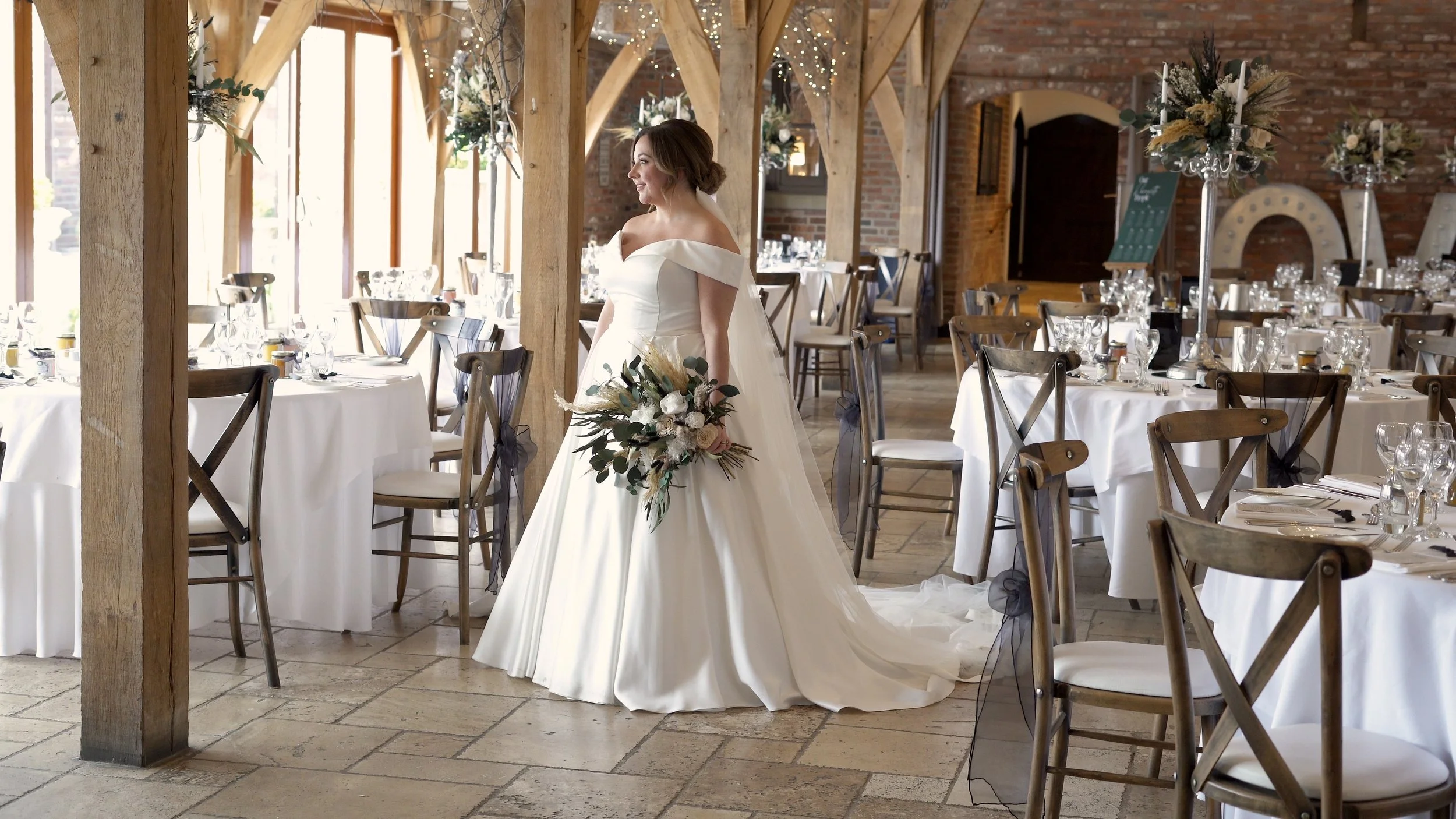 A bride in a white wedding gown holding a bouquet, standing in a decorated reception hall with round tables covered in white cloths, set with glassware, and surrounded by wooden chairs with black bows. The hall has wooden beams and brick walls, with 