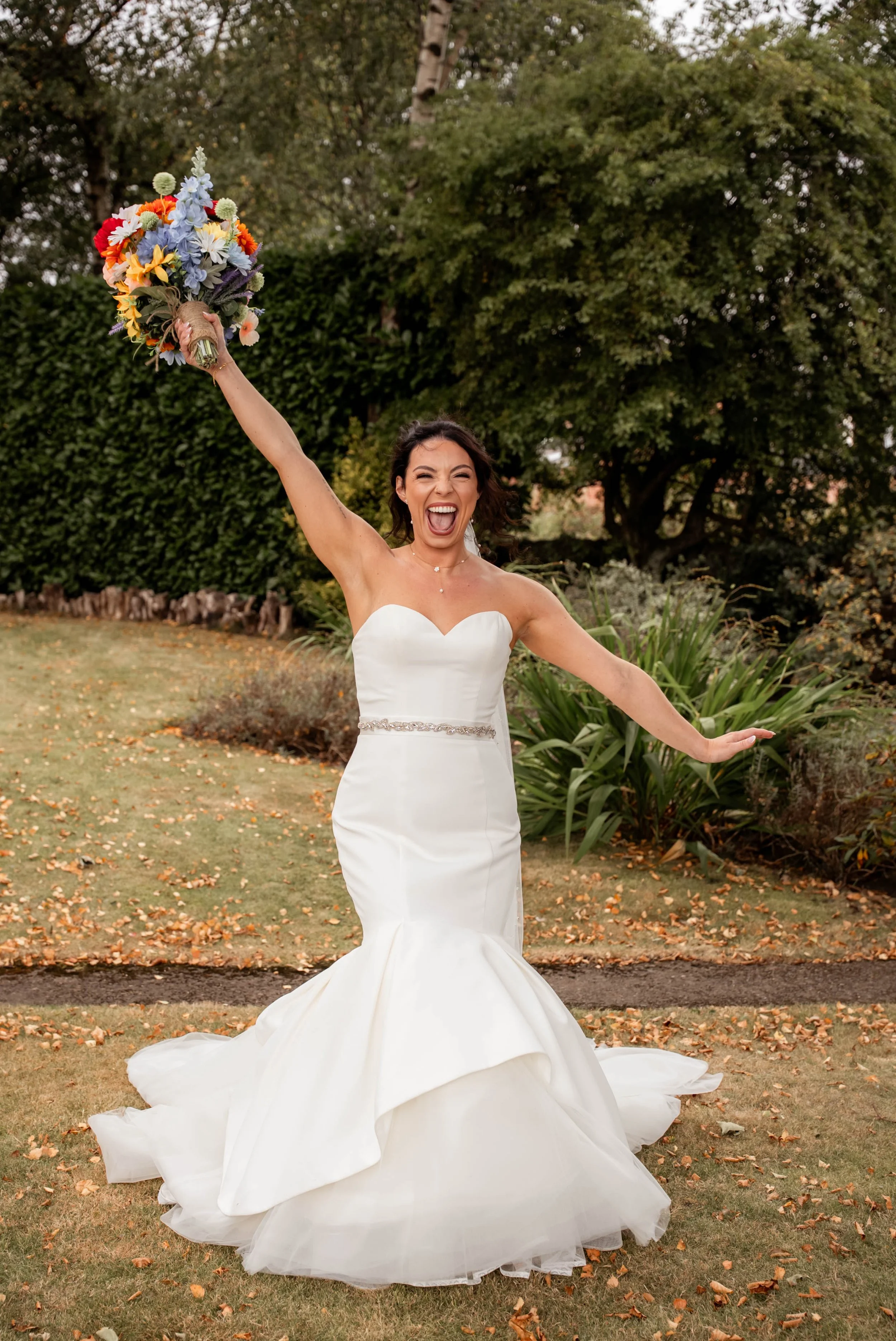 A joyful bride in a strapless white wedding gown with a fitted bodice and a mermaid skirt, holding a vibrant bouquet of flowers high in the air outdoors, surrounded by green trees and grass.