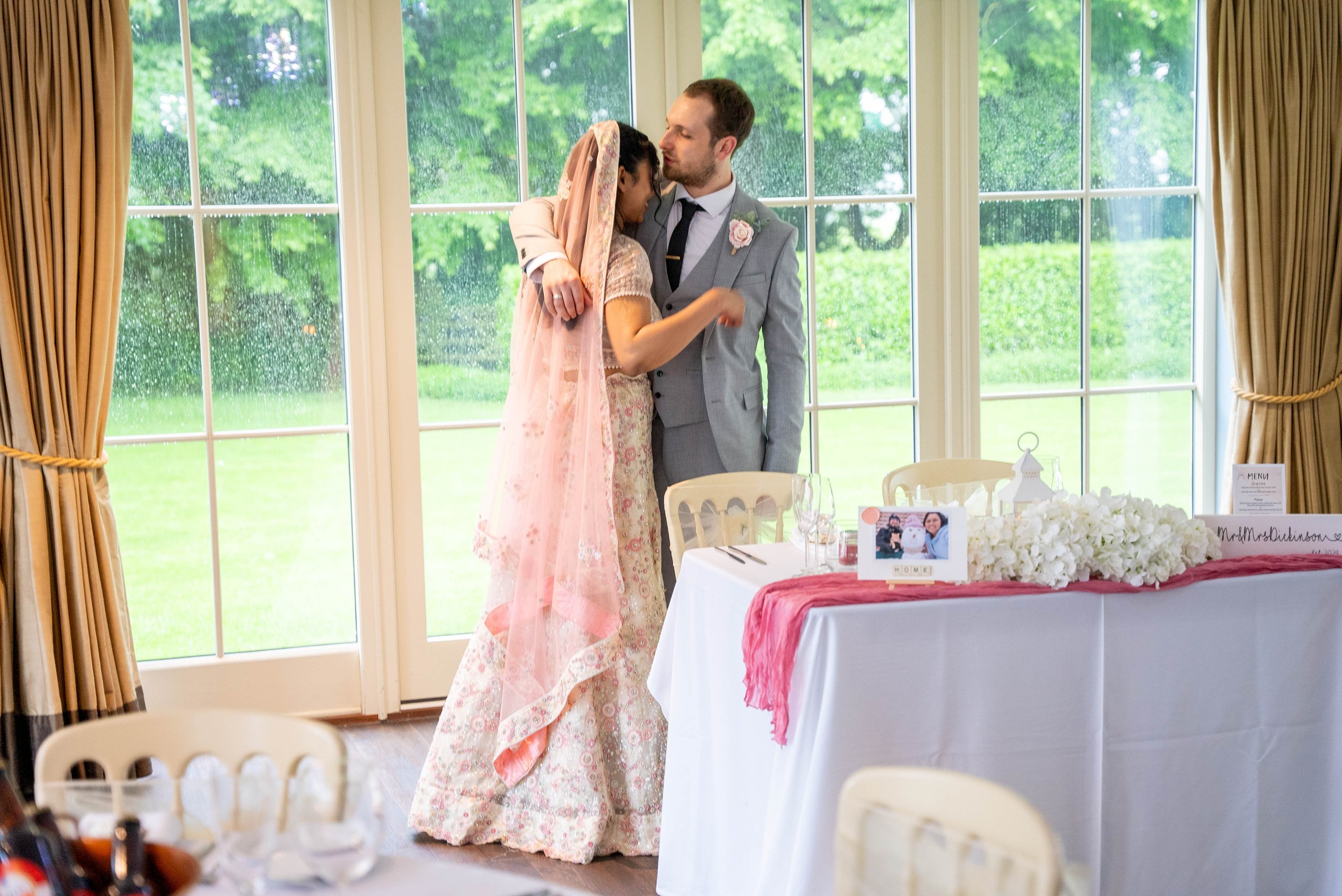 A bride and groom sharing a dance at their wedding reception inside a room with large windows and greenery outside. The bride wears a traditional dress with pink and gold embroidery and a pink veil. The groom is dressed in a light gray suit with a bl
