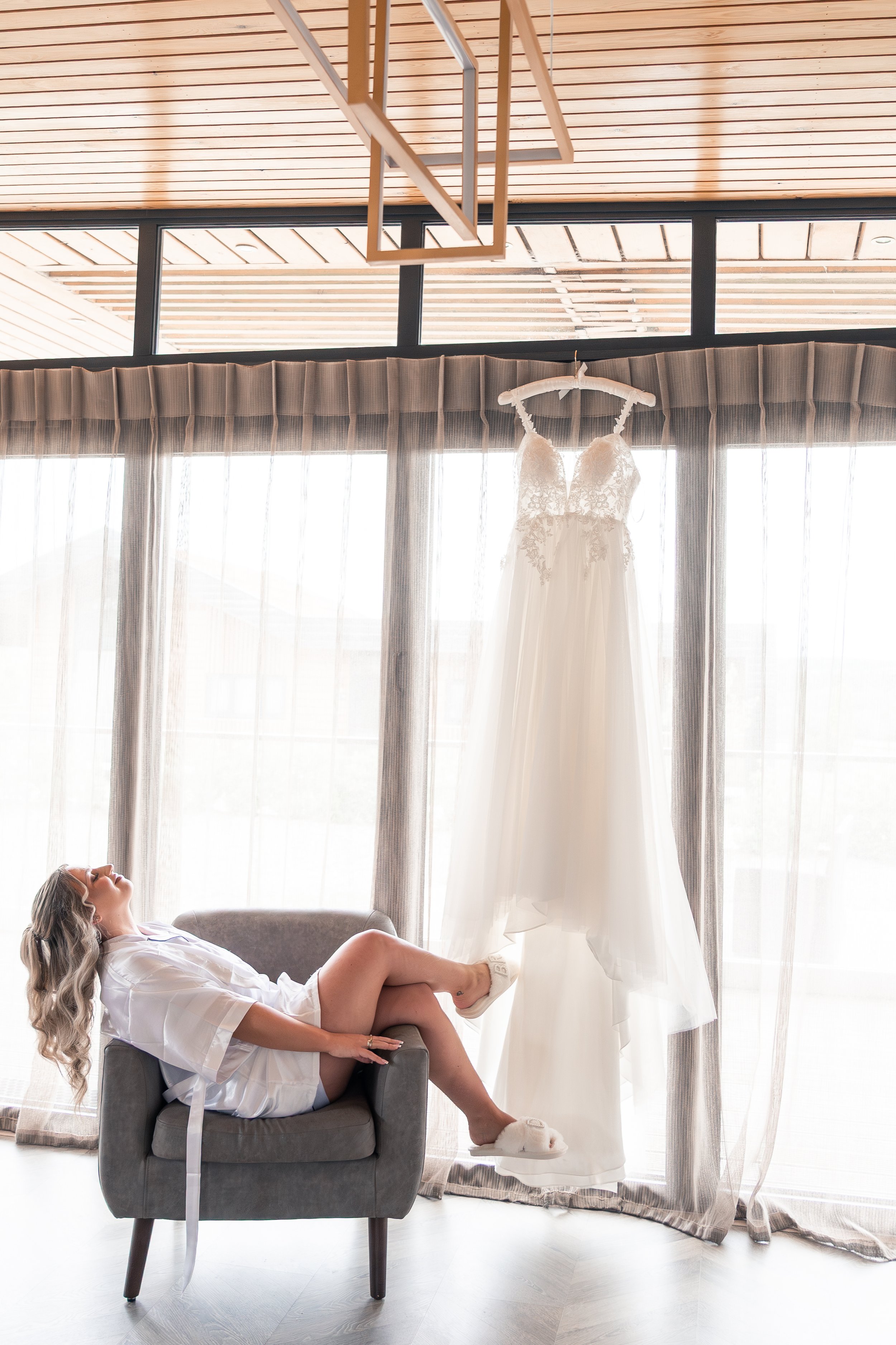 A woman relaxing on a gray armchair in front of a large window with light curtains, a wedding dress hanging from the curtain rod, and sunlight streaming in.