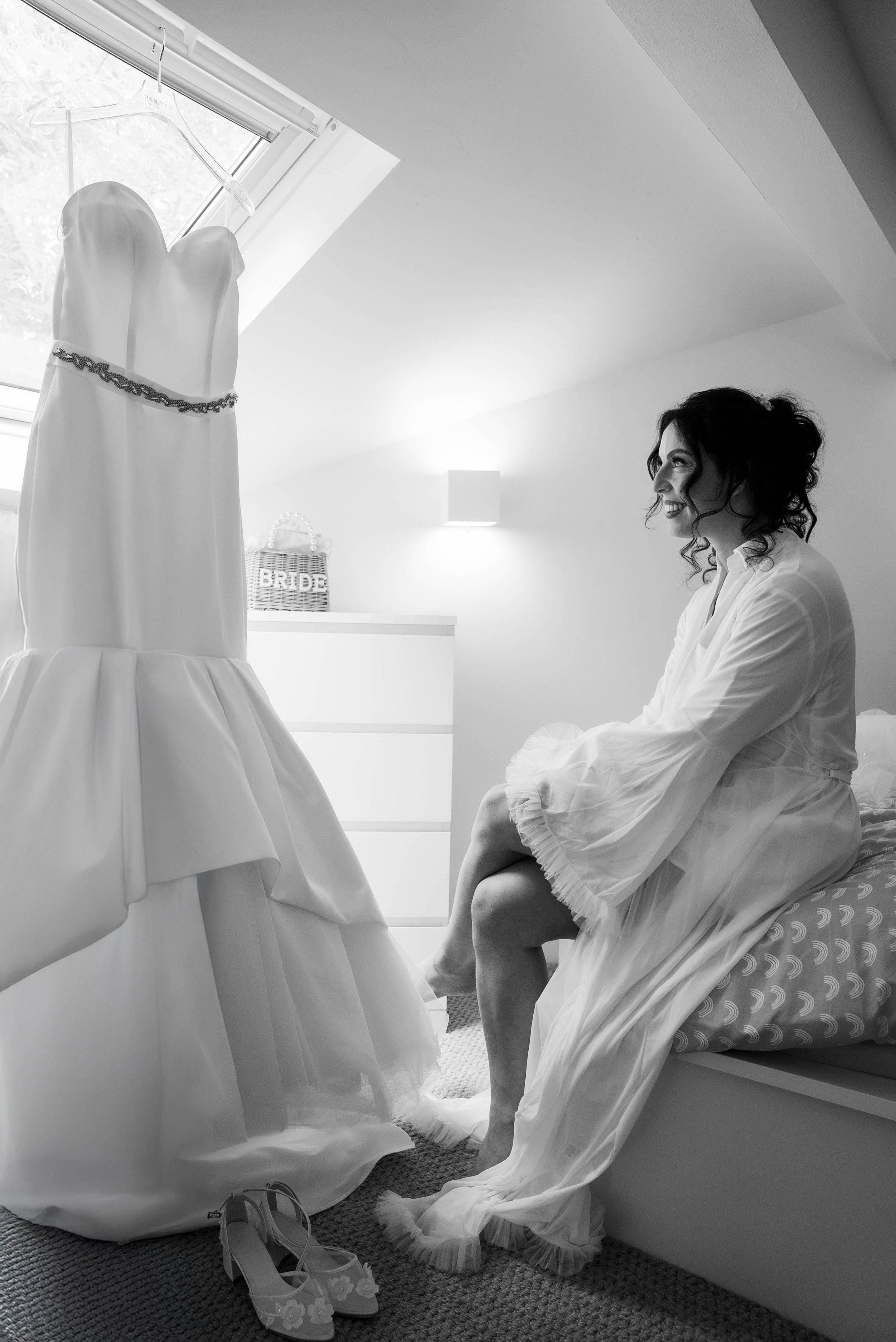 A woman in a robe sitting on a bed while looking at a wedding dress hanging near a window in a bright room.