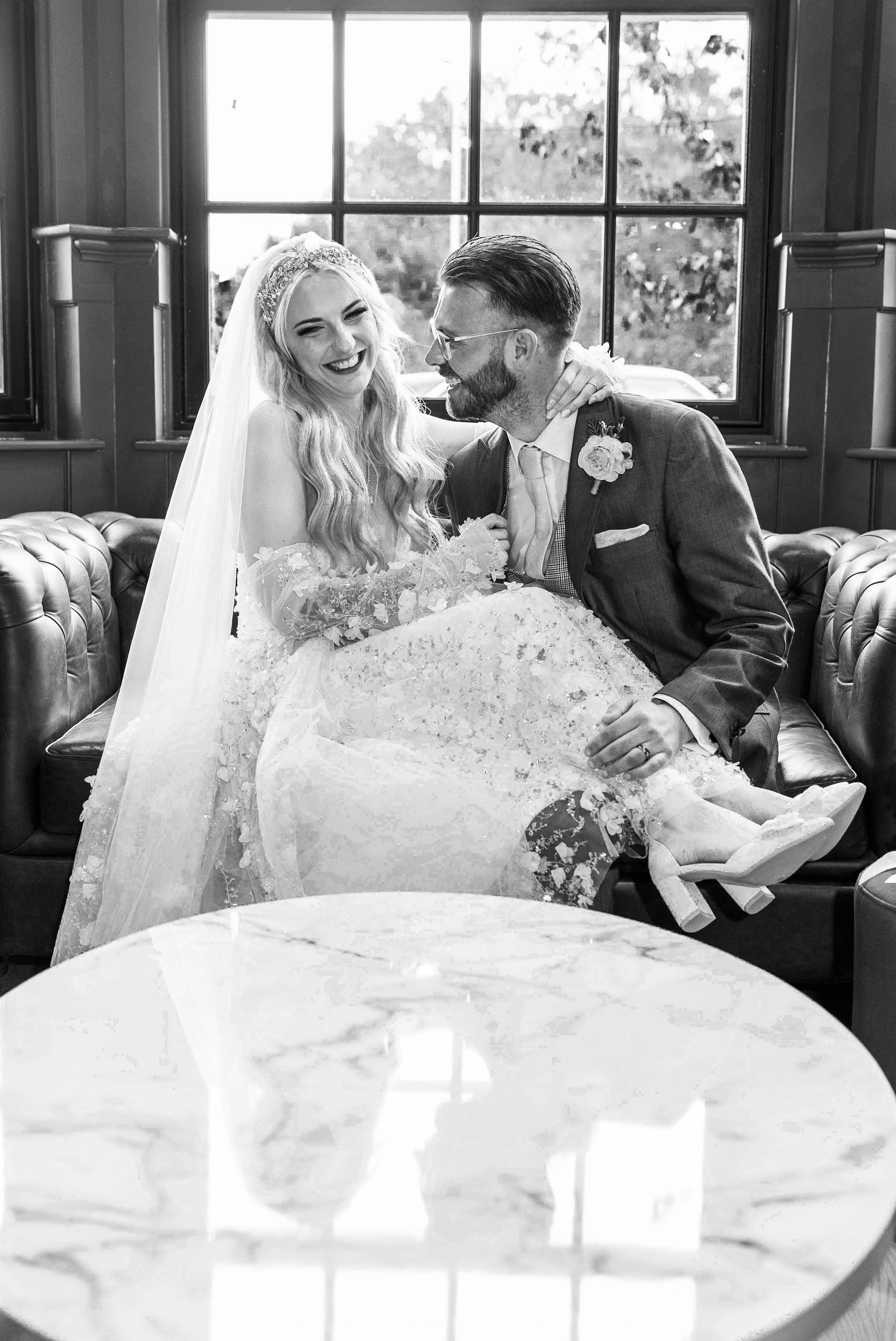 Black and white photo of a smiling bride in a wedding gown and veil, and a groom in glasses and a suit, sharing a joyful moment indoors by a large window.