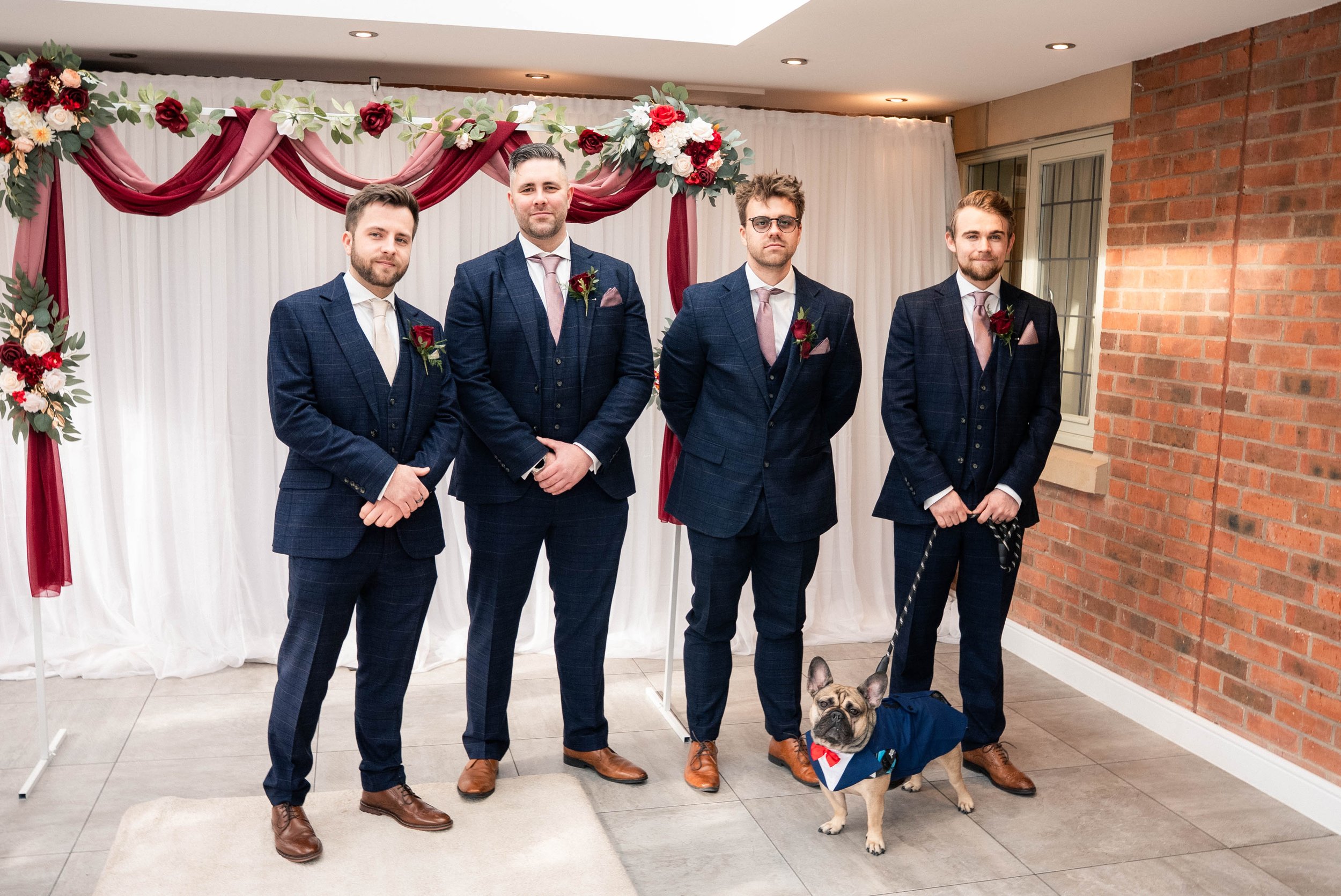 Four men dressed in dark blue suits with pink ties and boutonnières standing in front of a white curtain with red and white floral decorations, and a French Bulldog wearing a tuxedo costume in a room with a brick wall.