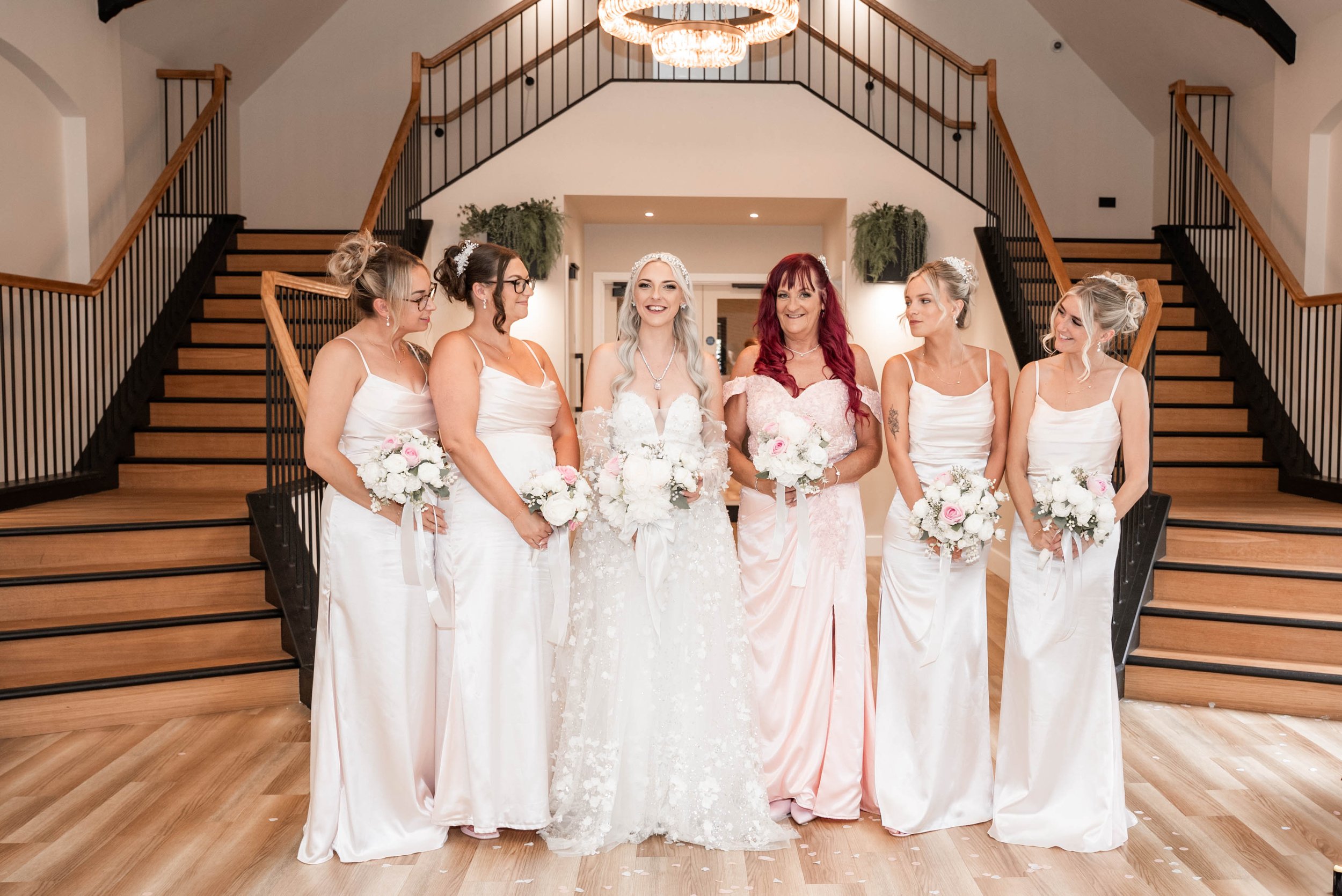 A group of six women, including the bride in a white lace wedding dress, standing in a ballroom with a chandelier and staircase in the background, holding bouquets of white and pink flowers.