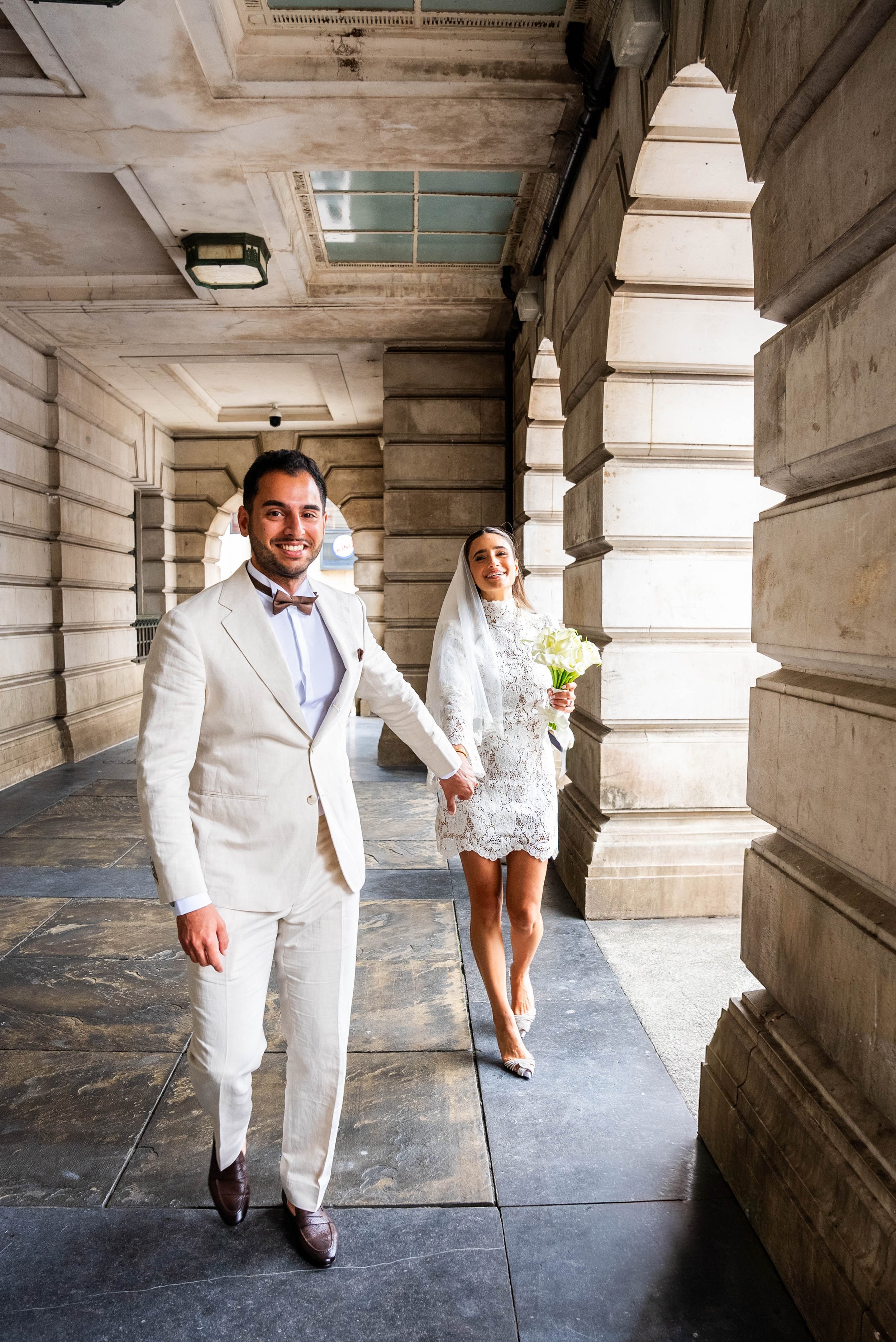 A newlywed couple holding hands and walking through a stone archway, the groom dressed in a cream suit and bowtie, and the bride in a lace dress holding a bouquet of white roses, all inside a historic stone building.