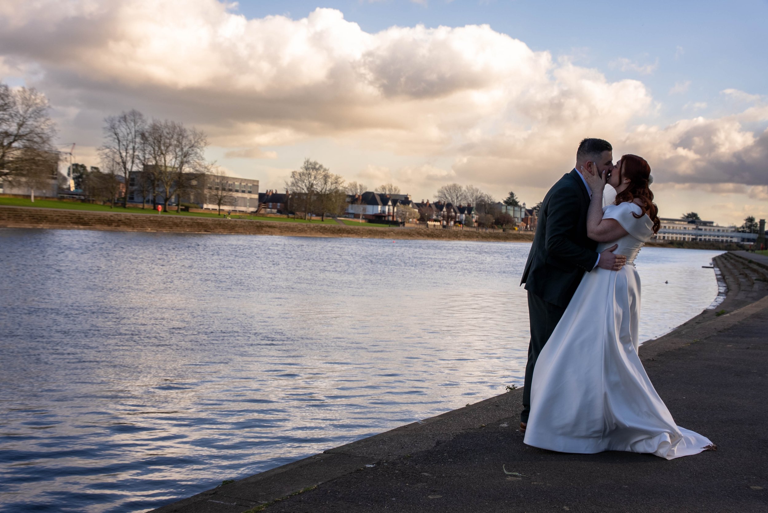 A bride and groom kissing by a river on a cloudy day.
