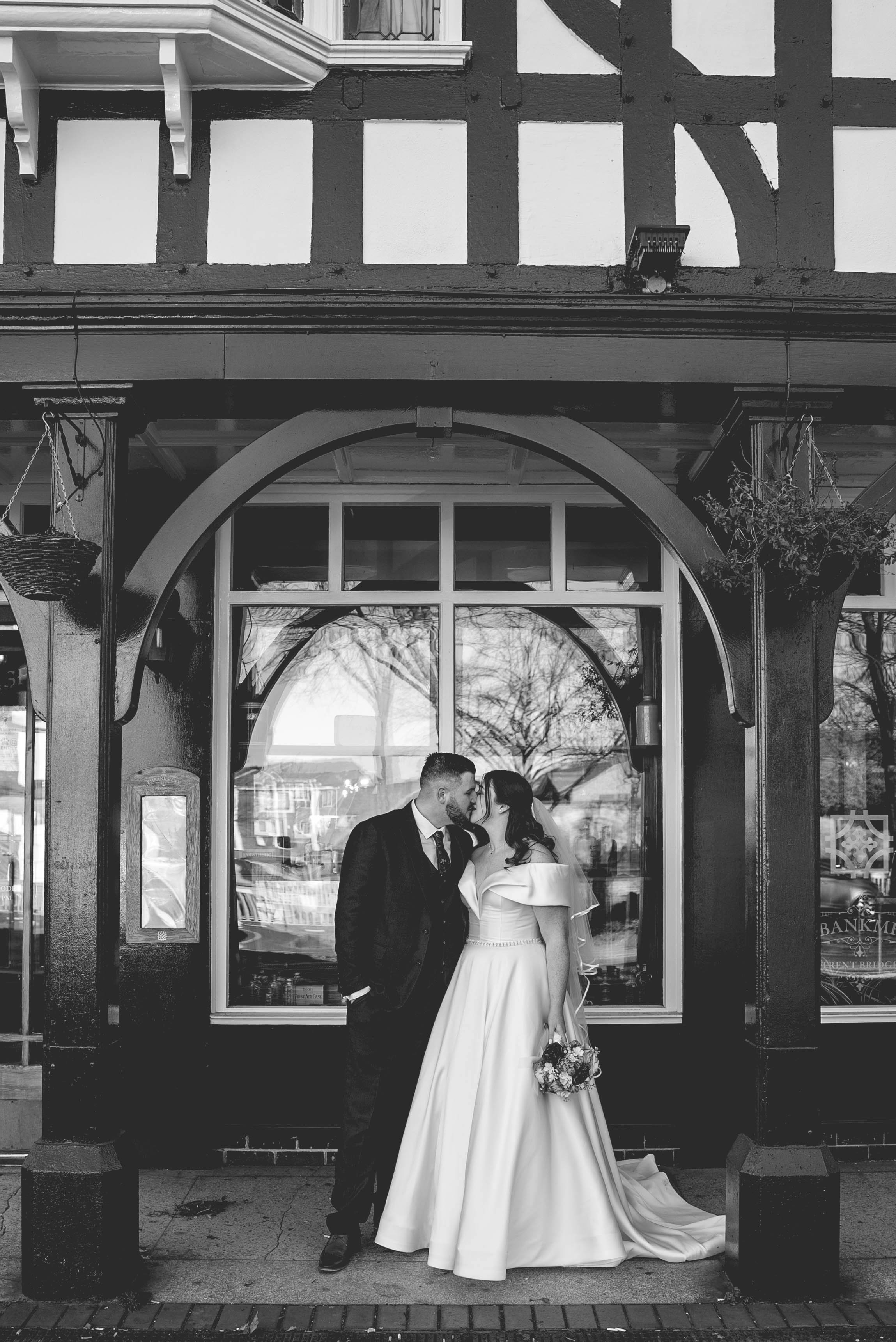 A black and white photo of a bride and groom standing close together outside a building with large windows. The couple is facing each other, with the groom in a dark suit and the bride in a long white dress holding a bouquet. They are about to kiss, 