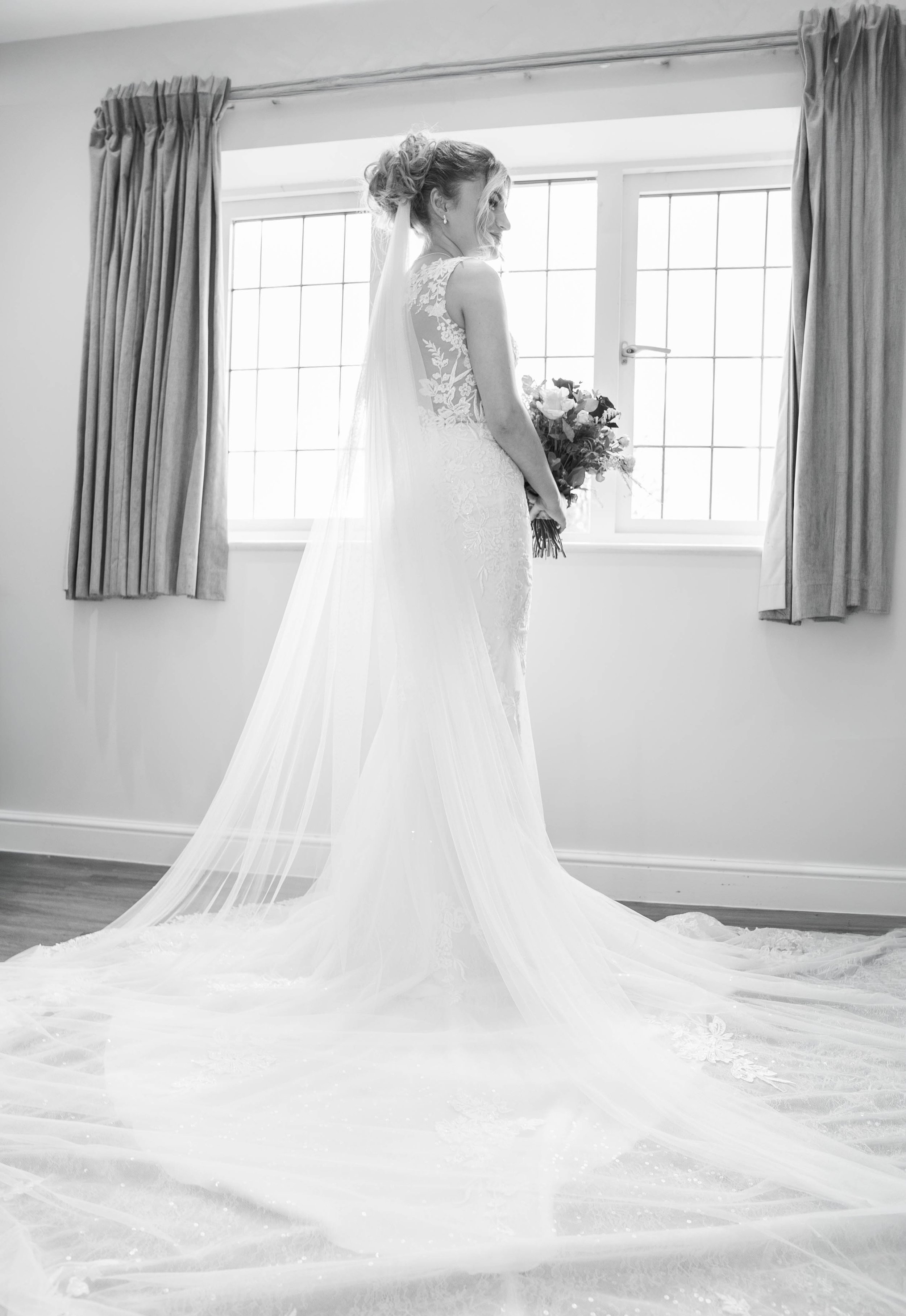 Bride in a wedding dress holding a bouquet, standing in front of a window, with curtains on either side.