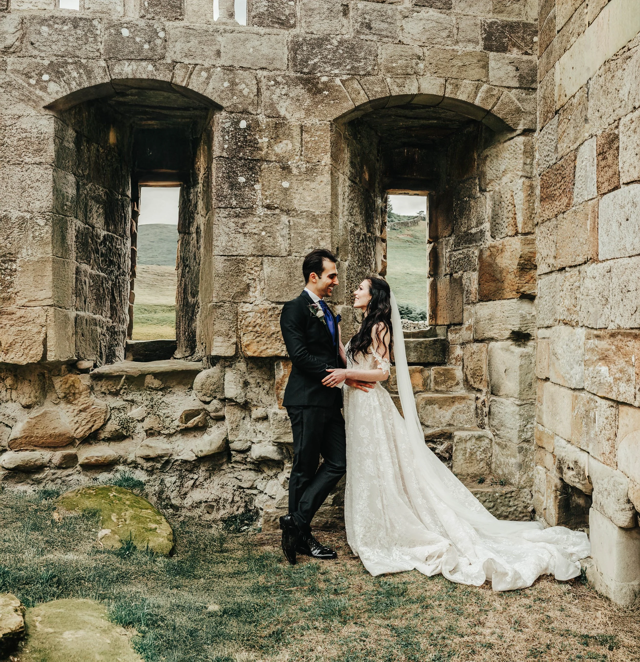 A bride and groom standing close together in front of an ancient stone structure with two arched windows, overlooking a green landscape.