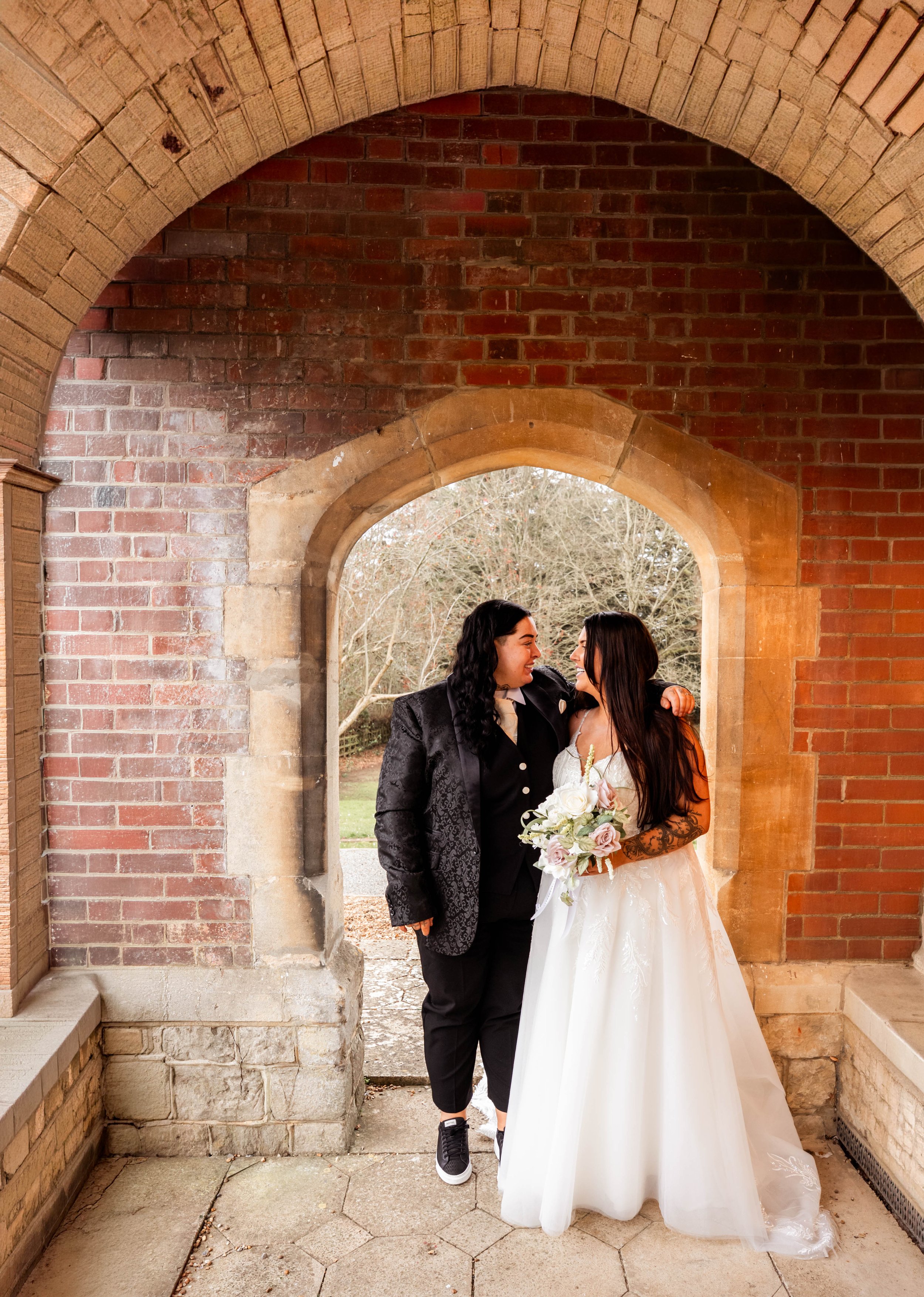 Two people, dressed in wedding attire, standing under an arched stone and brick structure outside, smiling at each other. The person on the left is wearing a black suit with a patterned blazer, and the person on the right is wearing a white wedding g