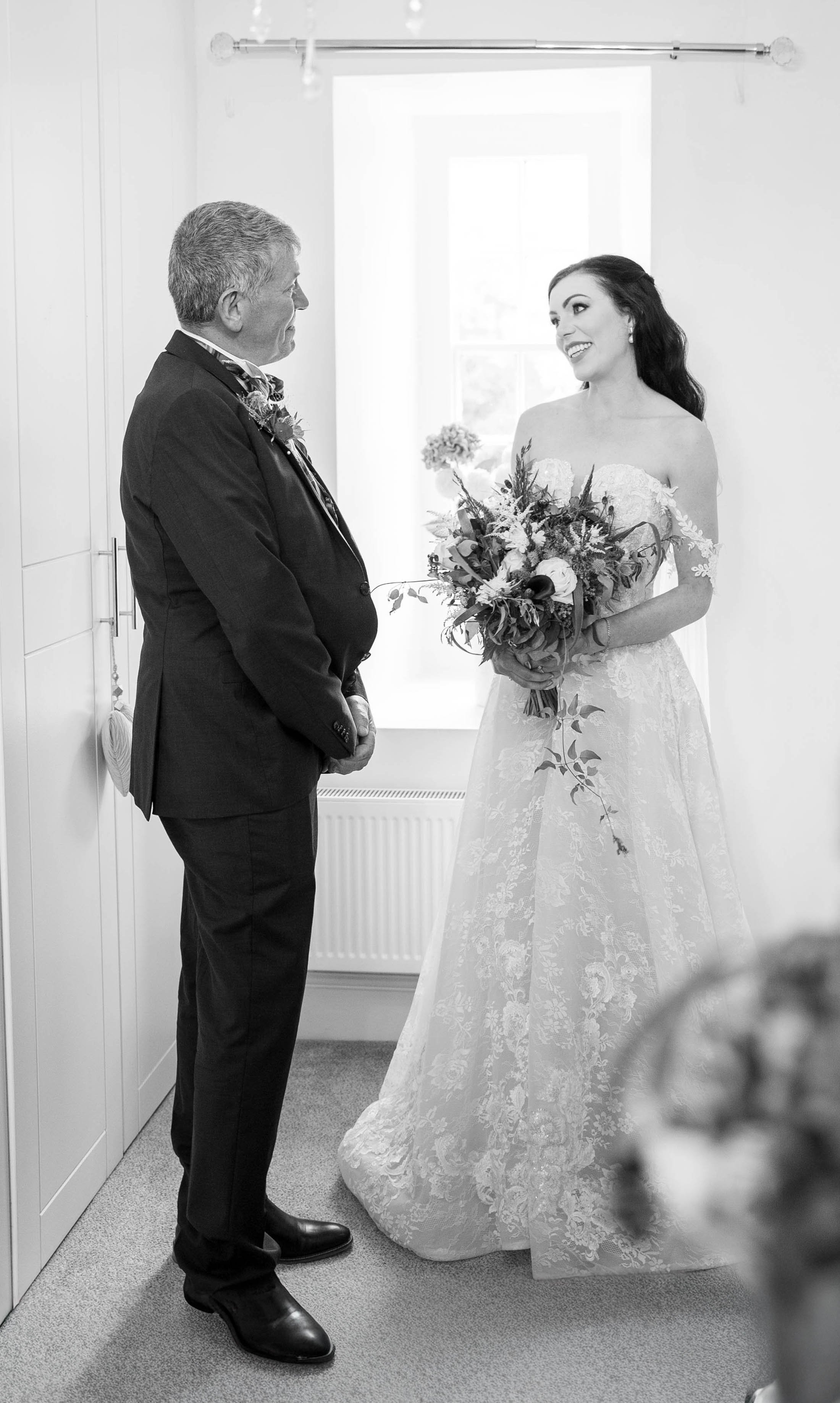 A bride holding a bouquet of flowers smiling at an older man, possibly her father, inside a bright room with a window in the background.