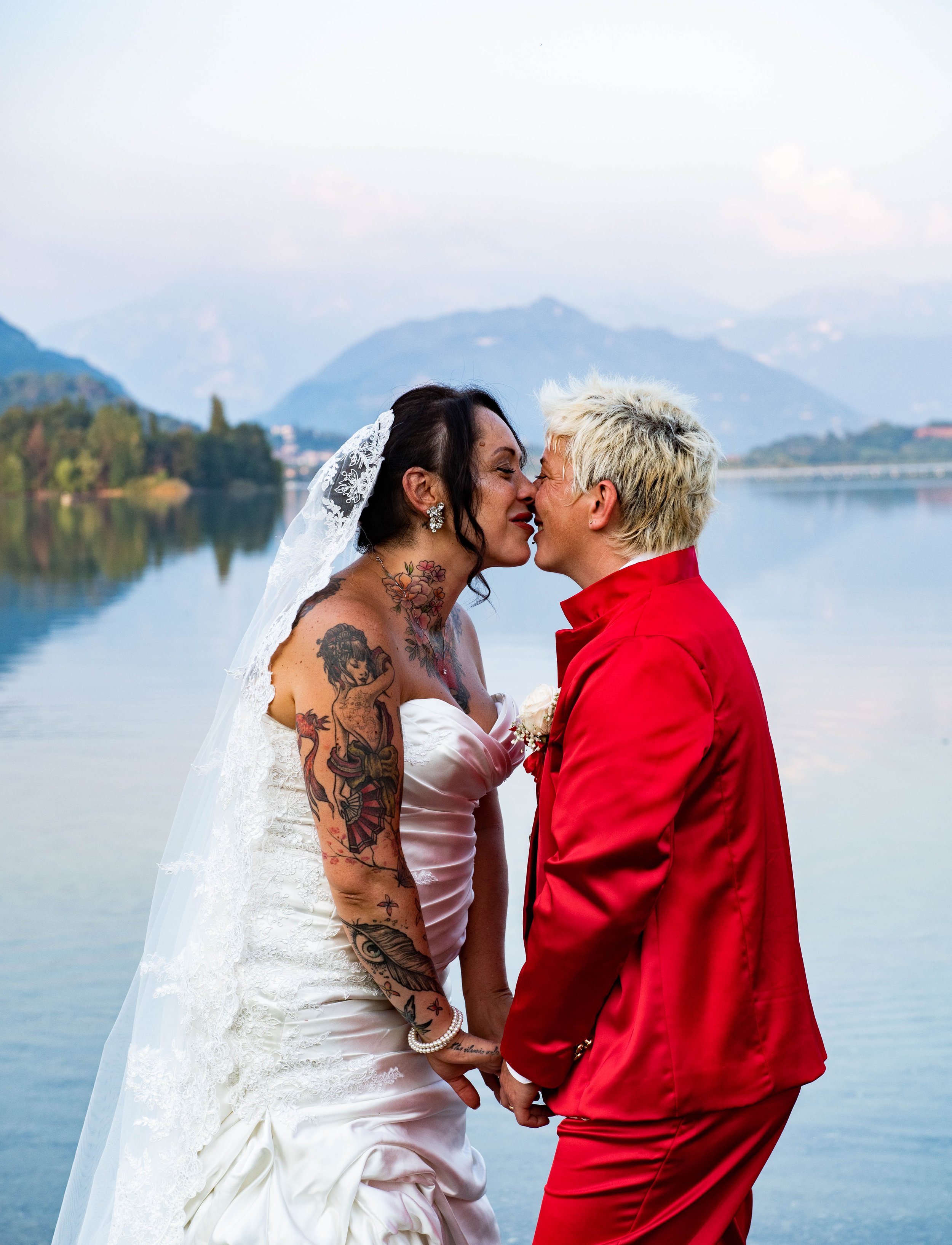 A same-sex couple in wedding attire kissing by a lake with mountains in the background.