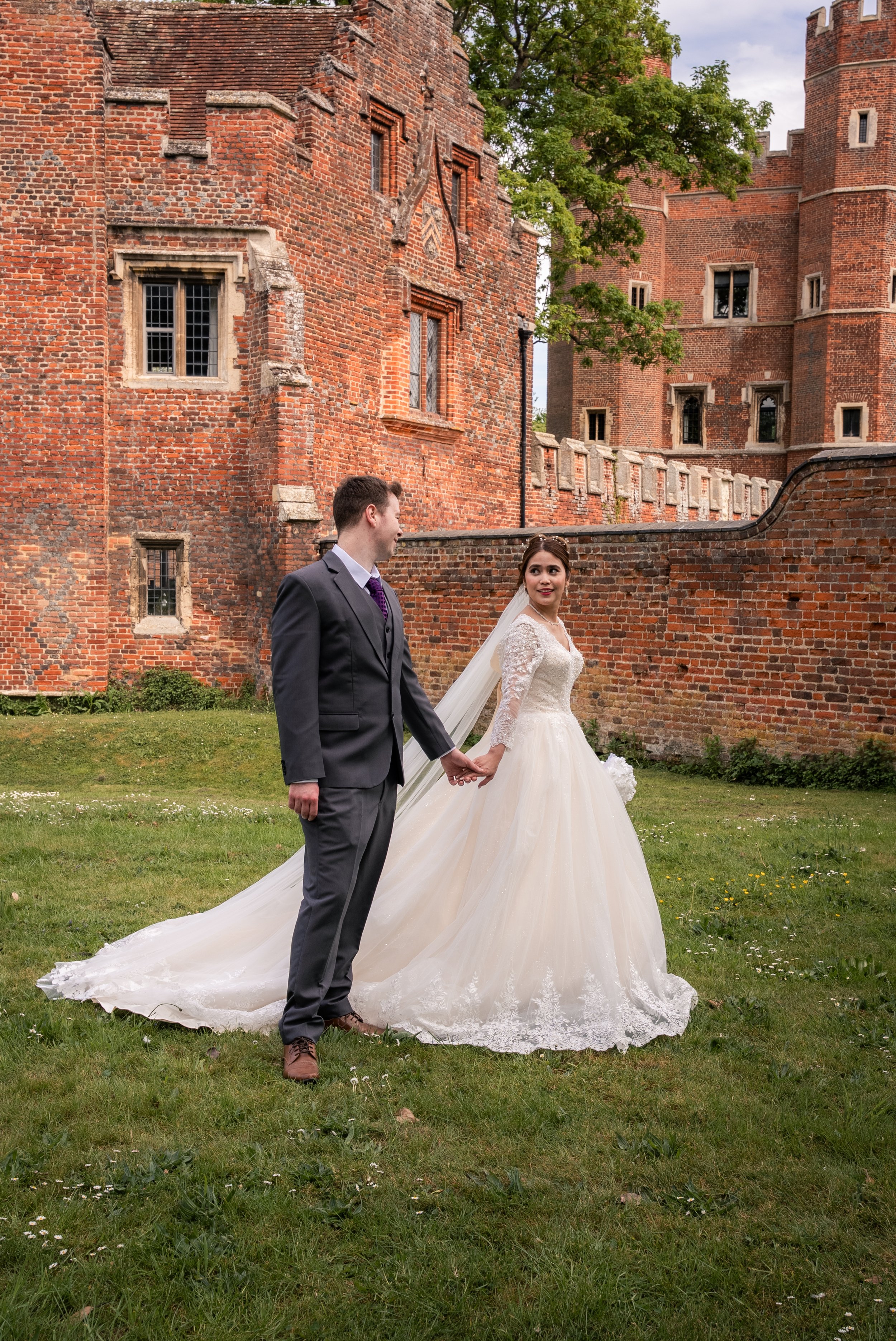 A bride and groom holding hands outside in front of a red brick castle, with the bride wearing a white wedding gown and veil, and the groom in a dark suit with a purple tie.