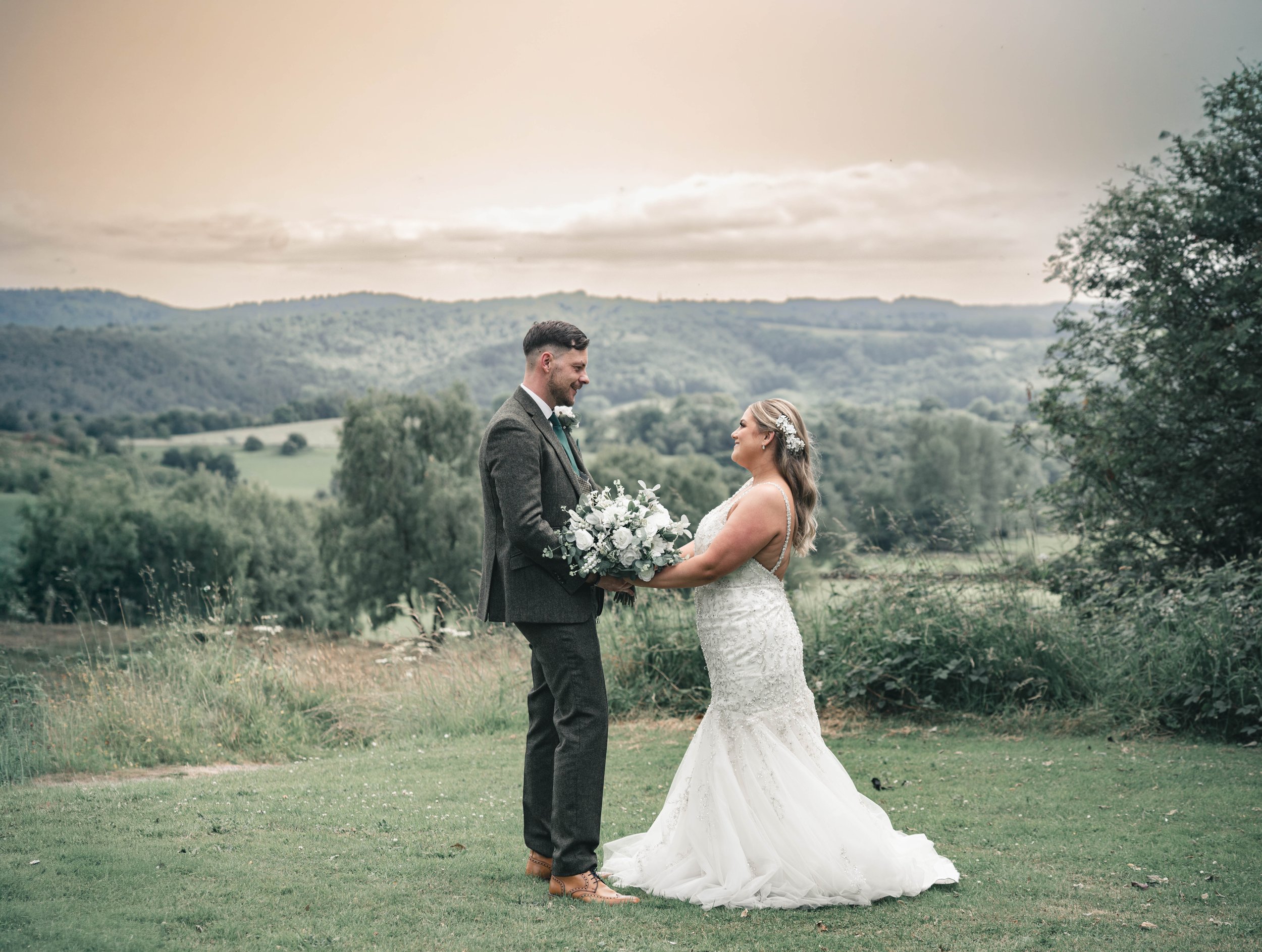 A wedding couple stands outdoors in a green field with rolling hills and trees in the background, facing each other and holding hands, with the bride in a white lace wedding gown and the groom in a gray suit.