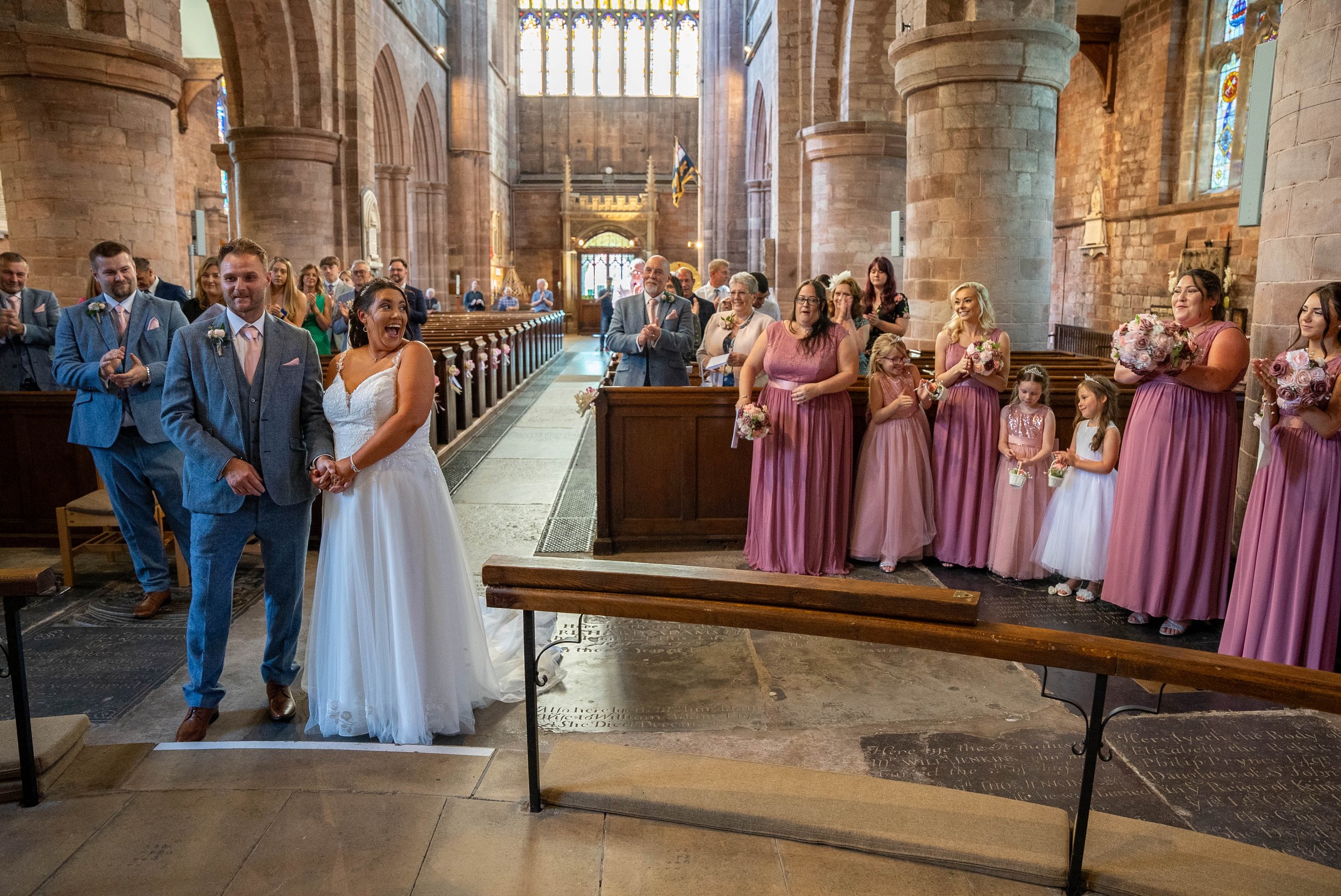 Bride and groom holding hands at the altar during a wedding ceremony inside a church, surrounded by bridesmaids and guests.