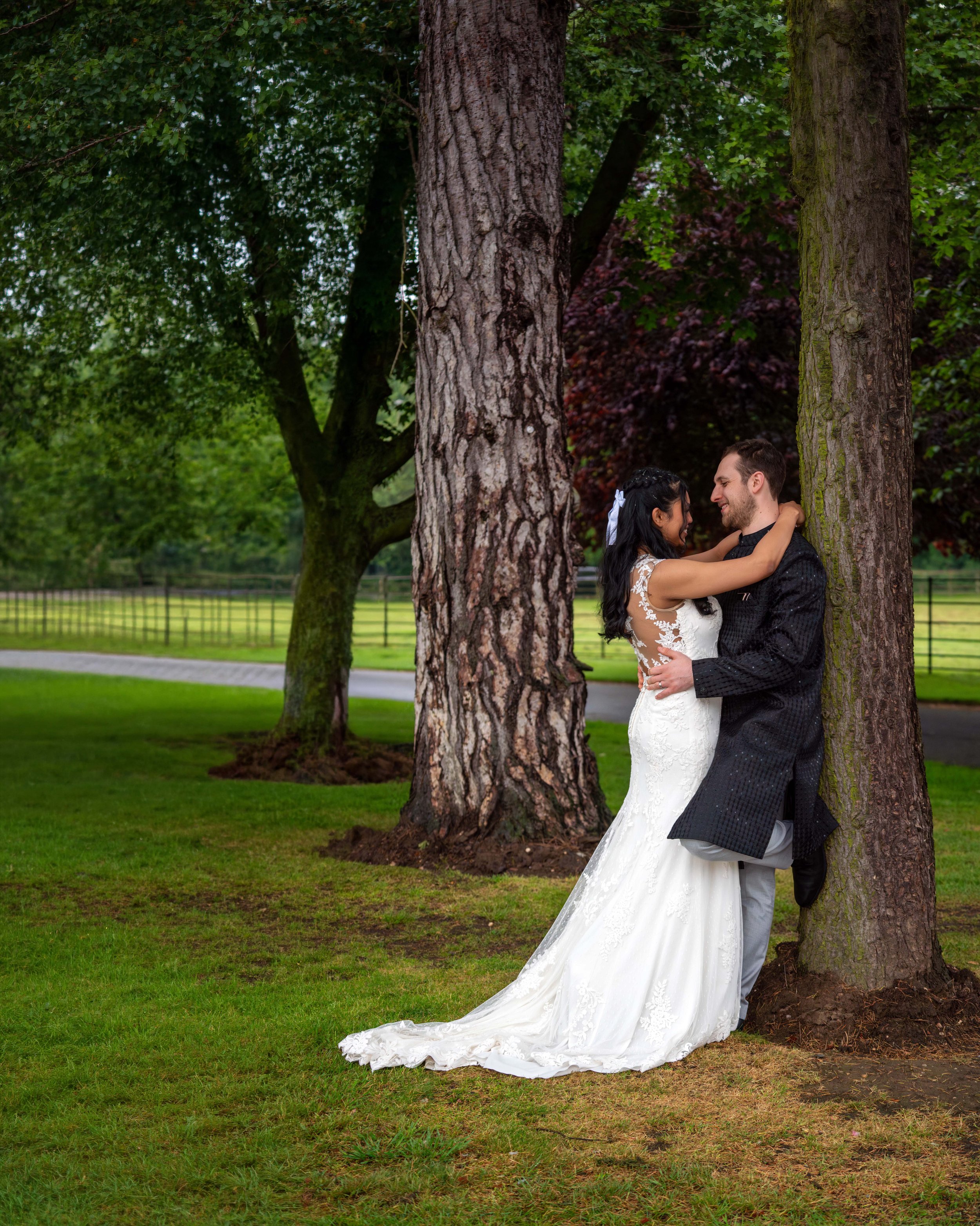 A newlywed couple in wedding attire embracing near a large tree in a park with lush green grass and trees in the background.