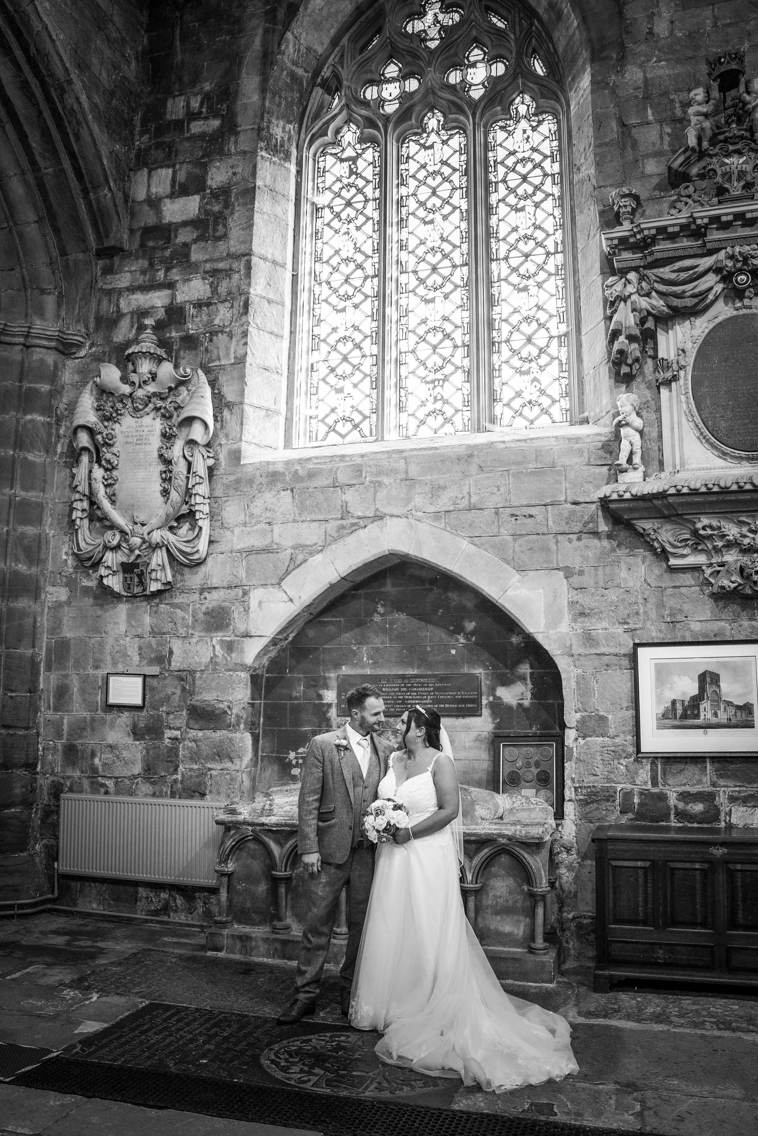 A bride and groom standing face-to-face inside a historic stone church, with stained glass windows overhead. The bride holds a bouquet, and they are smiling at each other.