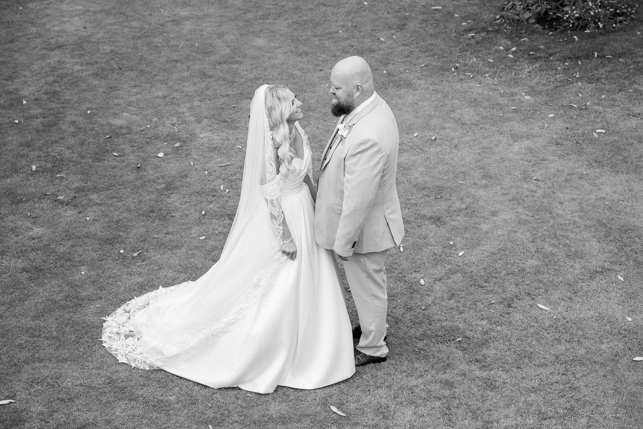 Black and white photo of a bride and groom standing on grass, looking at each other, outside.