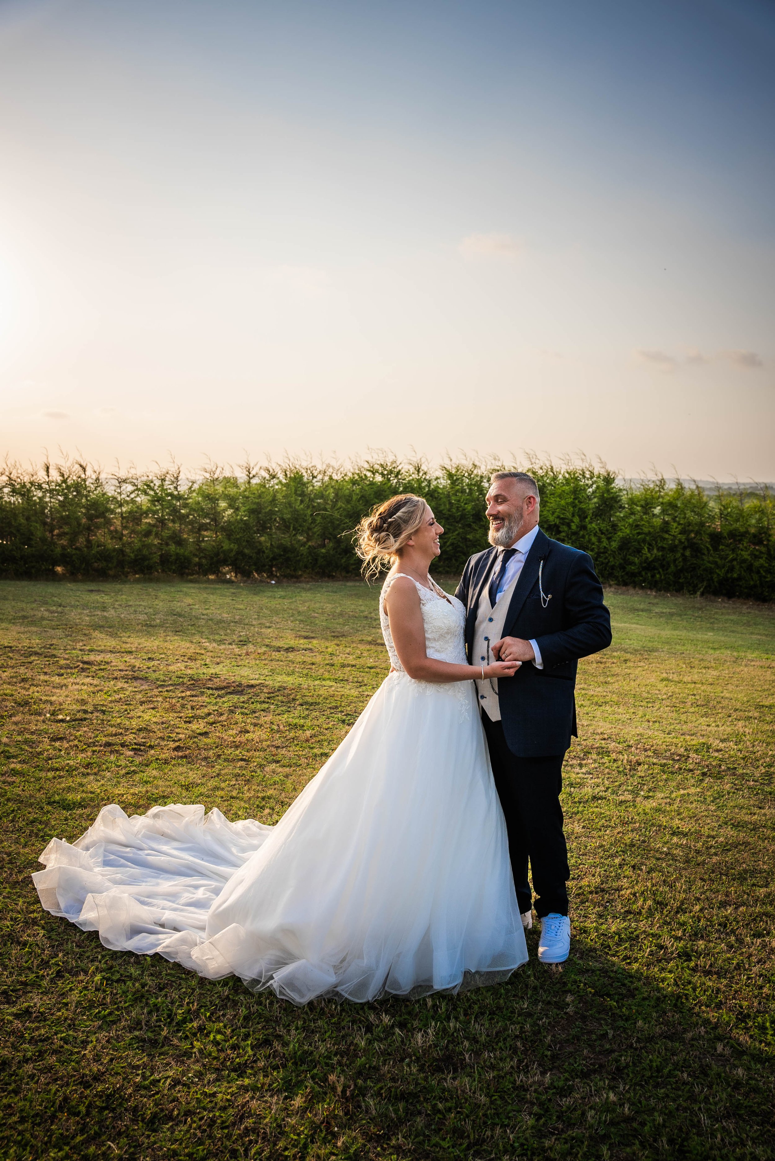 A bride and groom are holding hands and smiling at each other outdoors during sunset, with greenery and an open sky in the background. The bride wears a white wedding gown with a long train, and the groom wears a dark suit with a vest and dress shirt