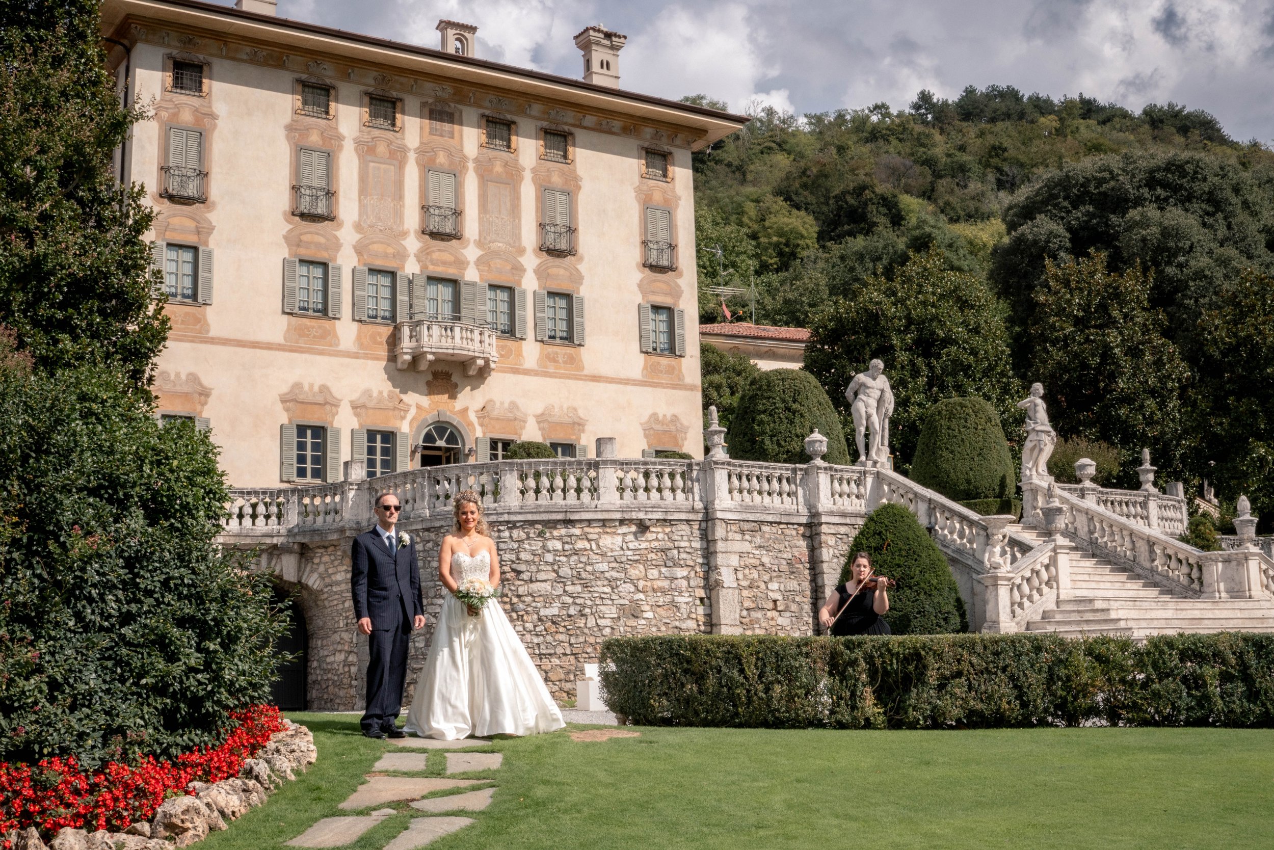 A wedding scene outside a large, ornate building with balconies and classical statues. The bride and groom are standing together, with the bride in a white wedding dress holding a bouquet, and the groom in a dark suit. A musician playing a violin is 