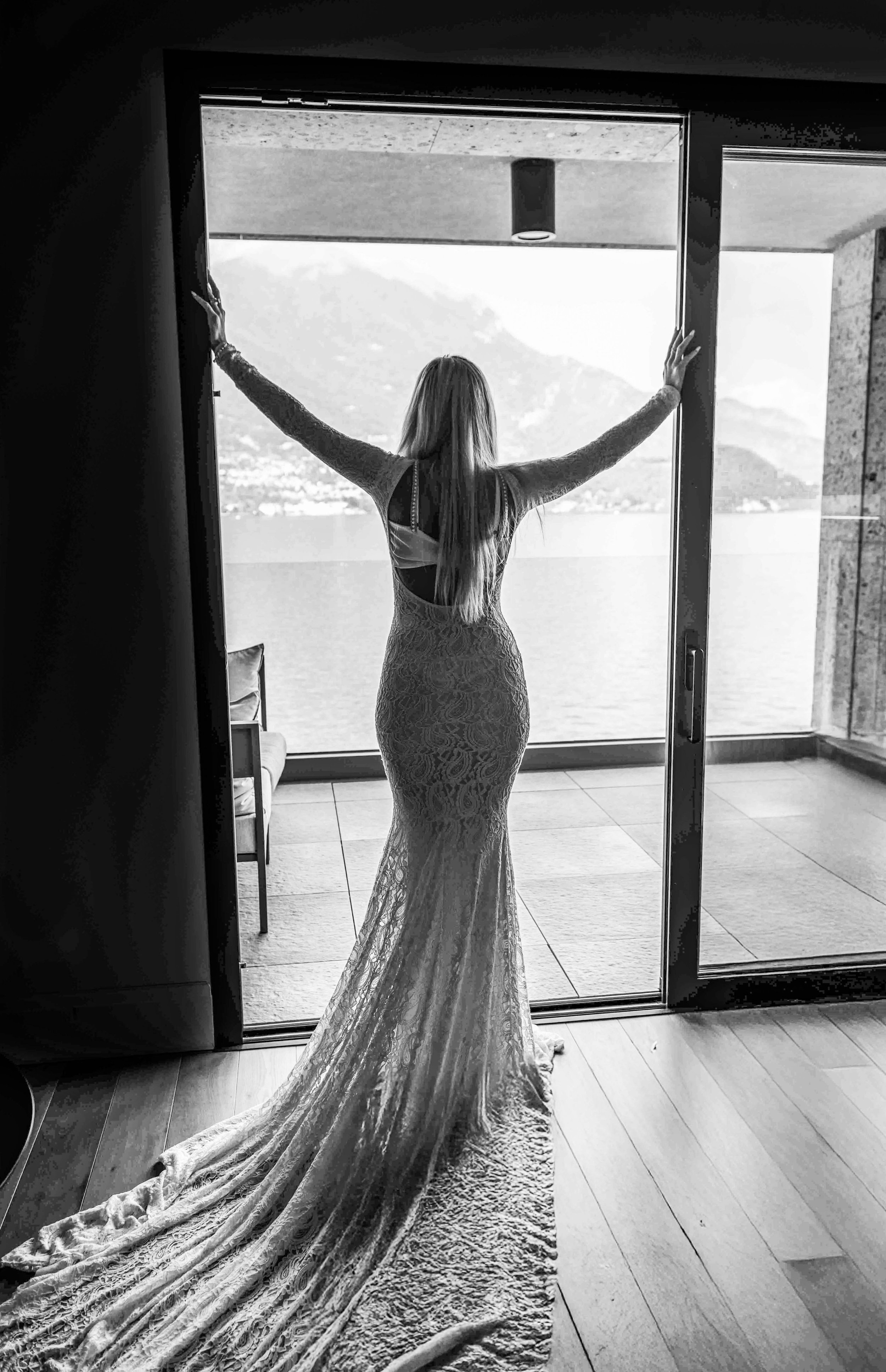 Woman in a lace wedding dress standing in front of a large window with her arms outstretched, overlooking a lake and mountains.
