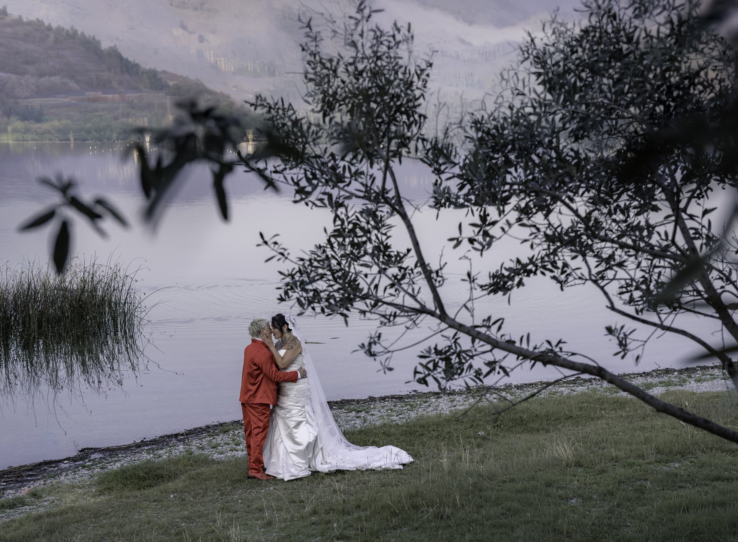 A wedding couple standing on the edge of a lake, embracing each other. The groom is dressed in a red suit, and the bride is wearing a white wedding gown with a long train. The scene is framed by trees and bushes, with mountains and a bridge visible i