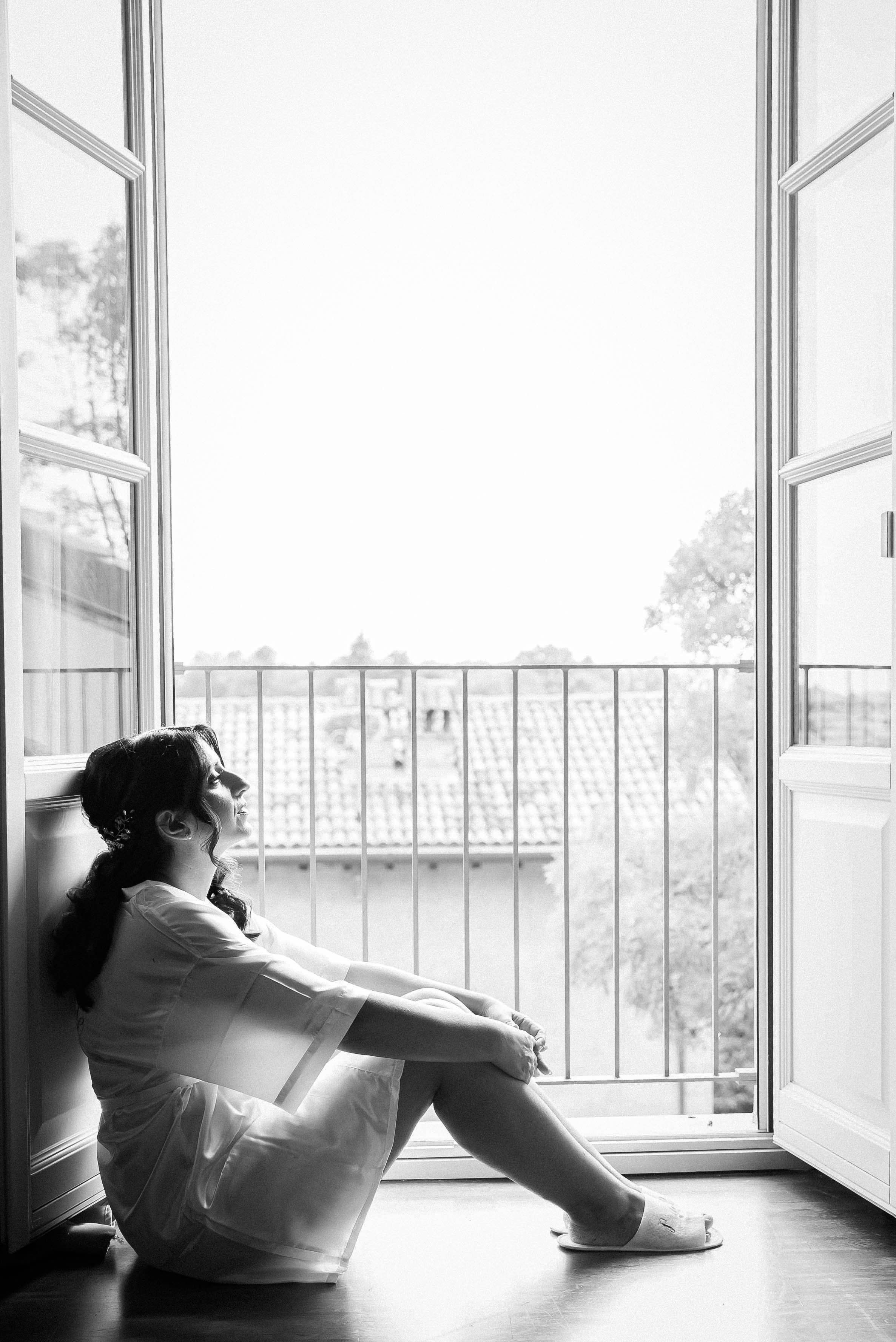 Black and white photo of a woman sitting by an open window with a view of trees and rooftops, relaxing with her legs extended and arms resting on her knees.