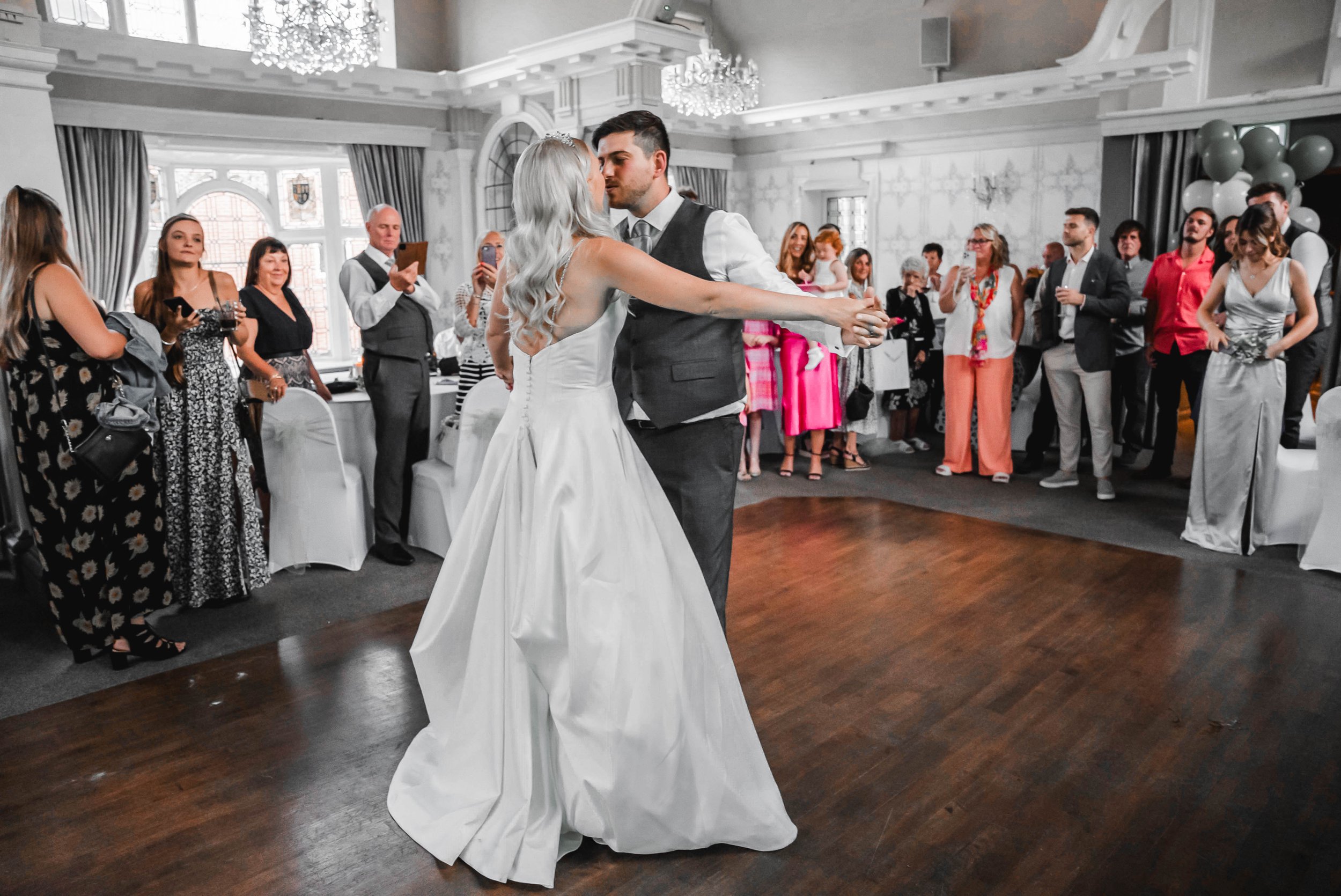 A bride and groom dance in the center of a wedding reception hall filled with guests watching and taking photos.