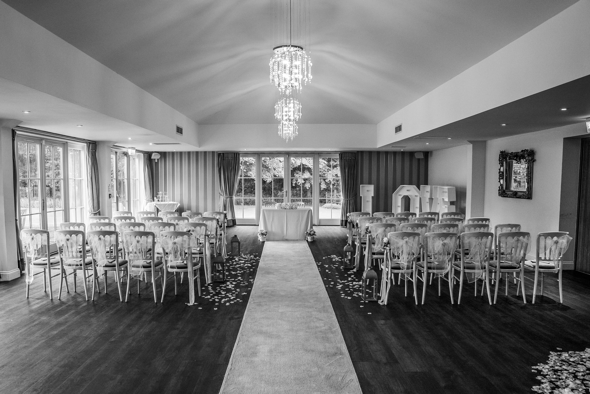 A decorated wedding ceremony space with rows of chairs on either side of a central aisle, adorned with tulle bows and scattered flower petals, leading to an altar with large decorative letters spelling 'LOVE' and large windows in the background.