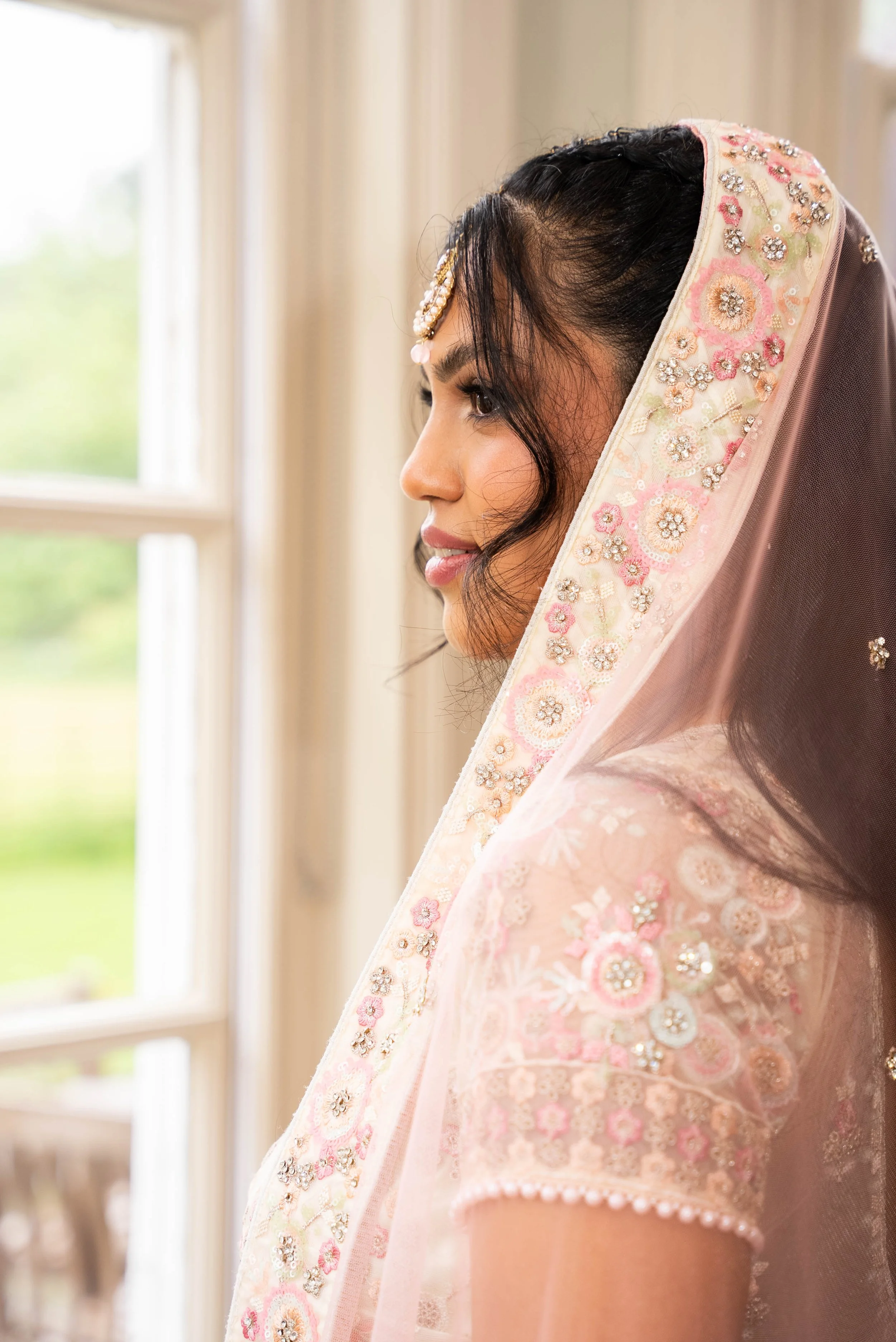 A woman dressed in traditional South Asian attire, wearing a pink embroidered saree with a matching veil, standing by a window with greenery outside.
