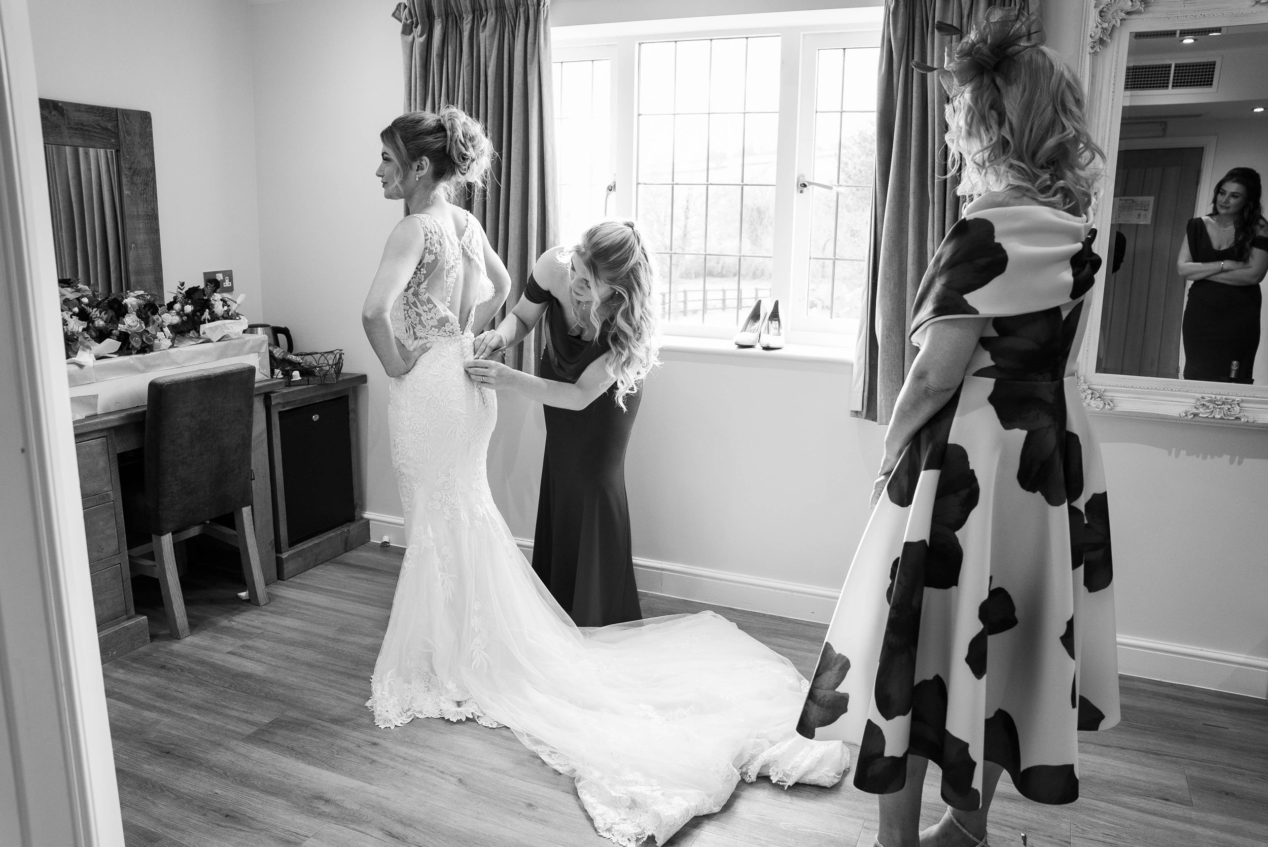 A bride getting help with her wedding dress from a woman in a dark gown as another woman in a floral dress watches in a bright room with a window, a mirror, and shoes on the windowsill.