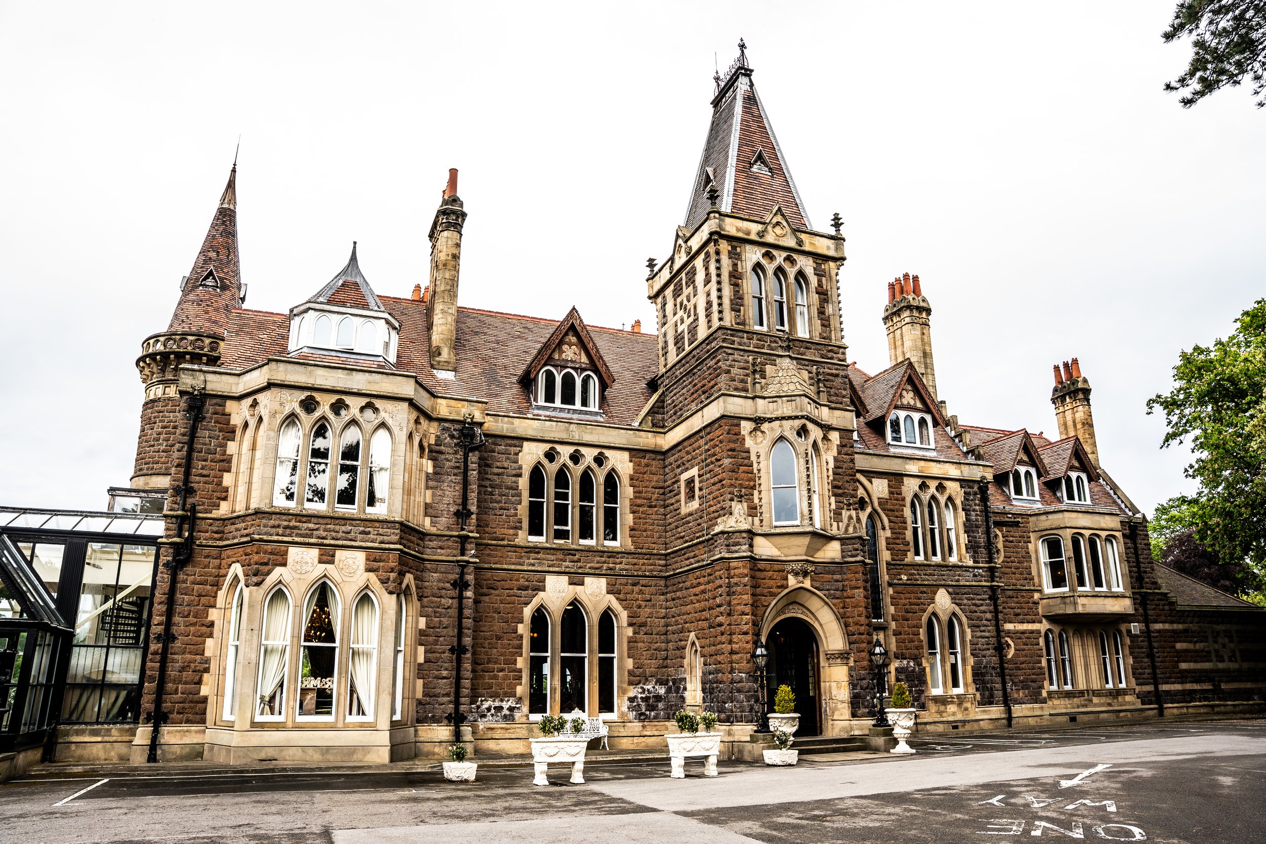 A historic stone building with gothic architectural features, including pointed arches and tall towers topped with steep spires, surrounded by a parking lot.