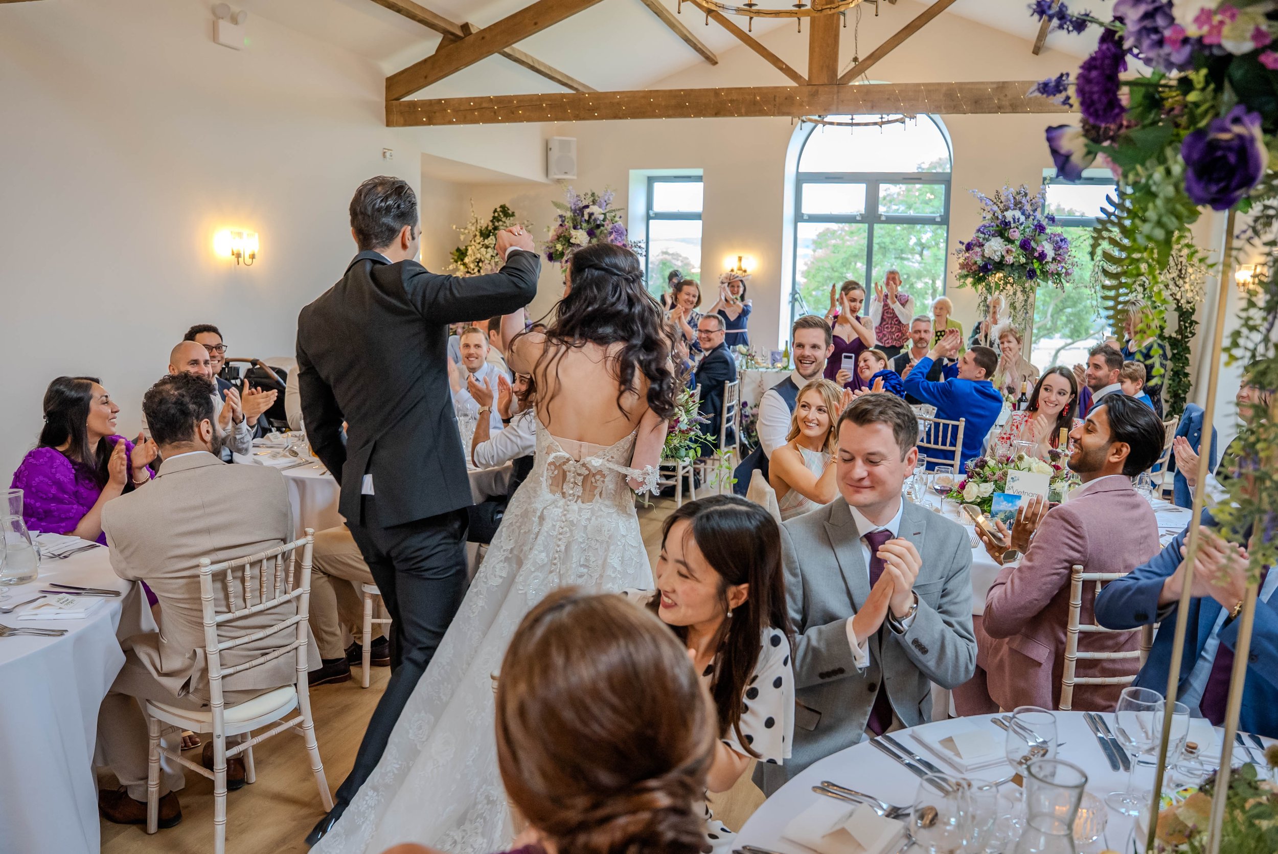 A wedding reception with a bride and groom dancing in the center while guests sit and watch, clapping and laughing, in a decorated hall with large windows and floral arrangements.
