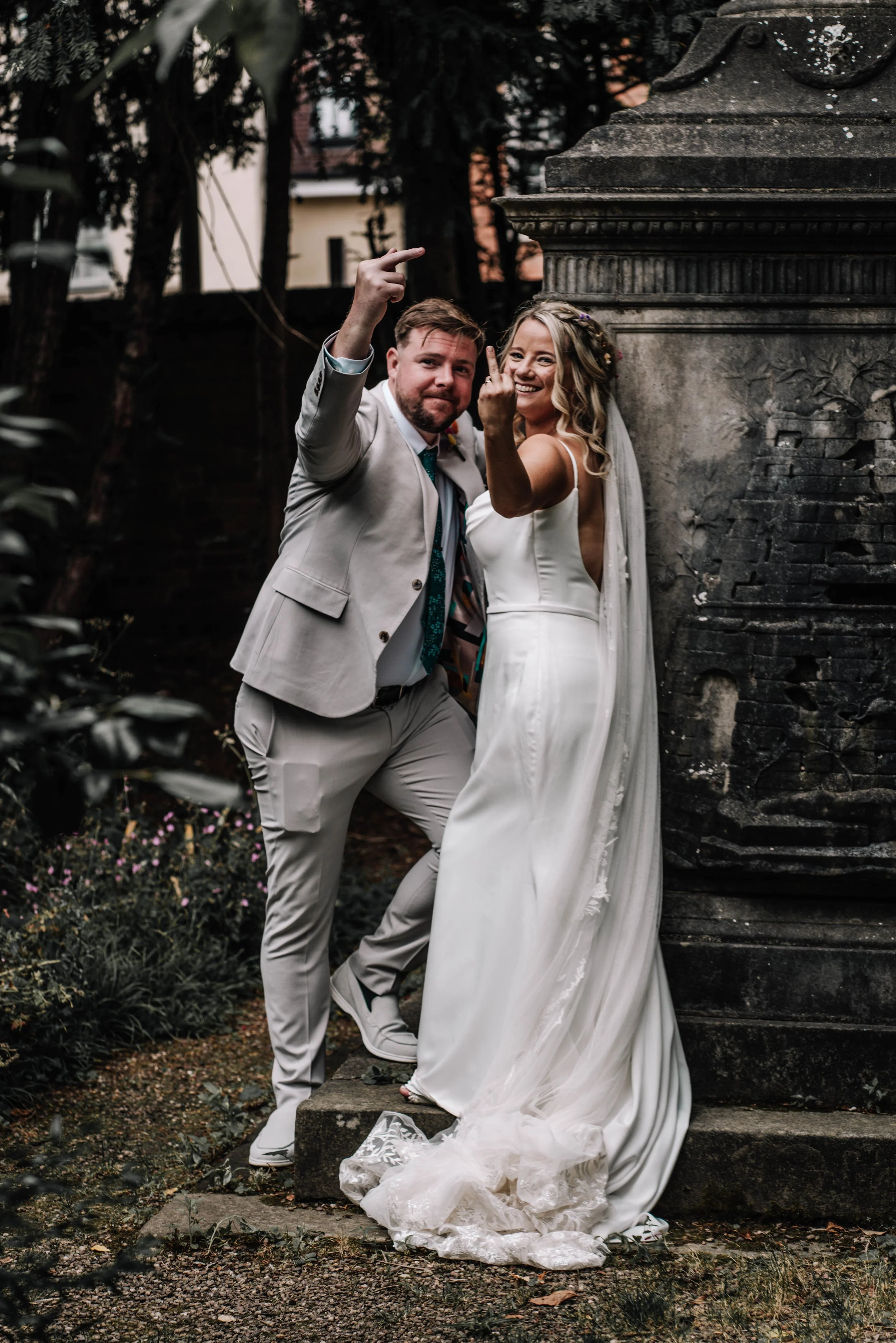 A bride and groom in wedding attire posing playfully outdoors near a stone monument with trees and buildings in the background.