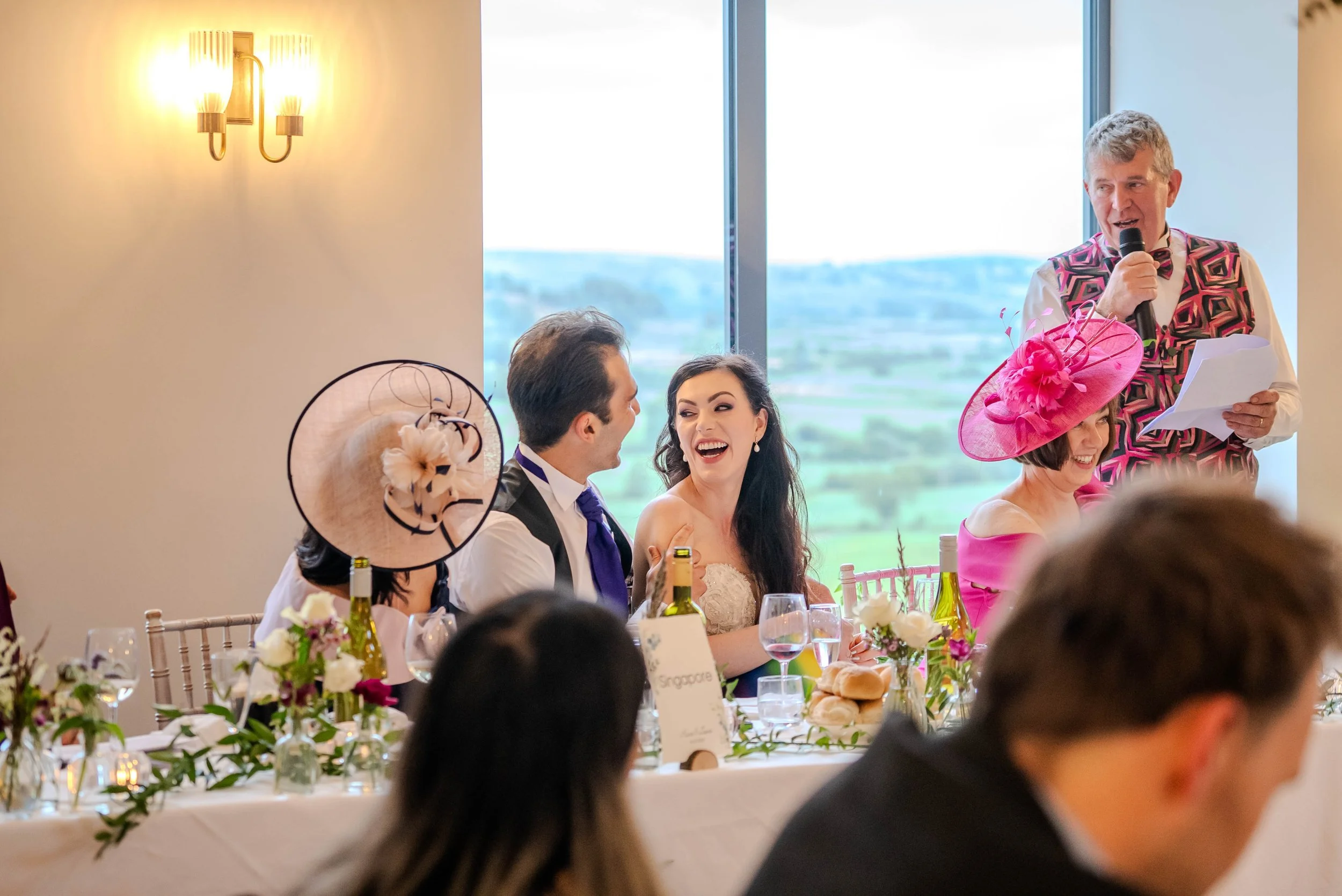 A newlywed couple at their wedding reception, sitting at a decorated banquet table with floral arrangements and wine, smiling and looking at each other. An older woman in a fancy pink dress and large pink hat and a man standing with a microphone are 