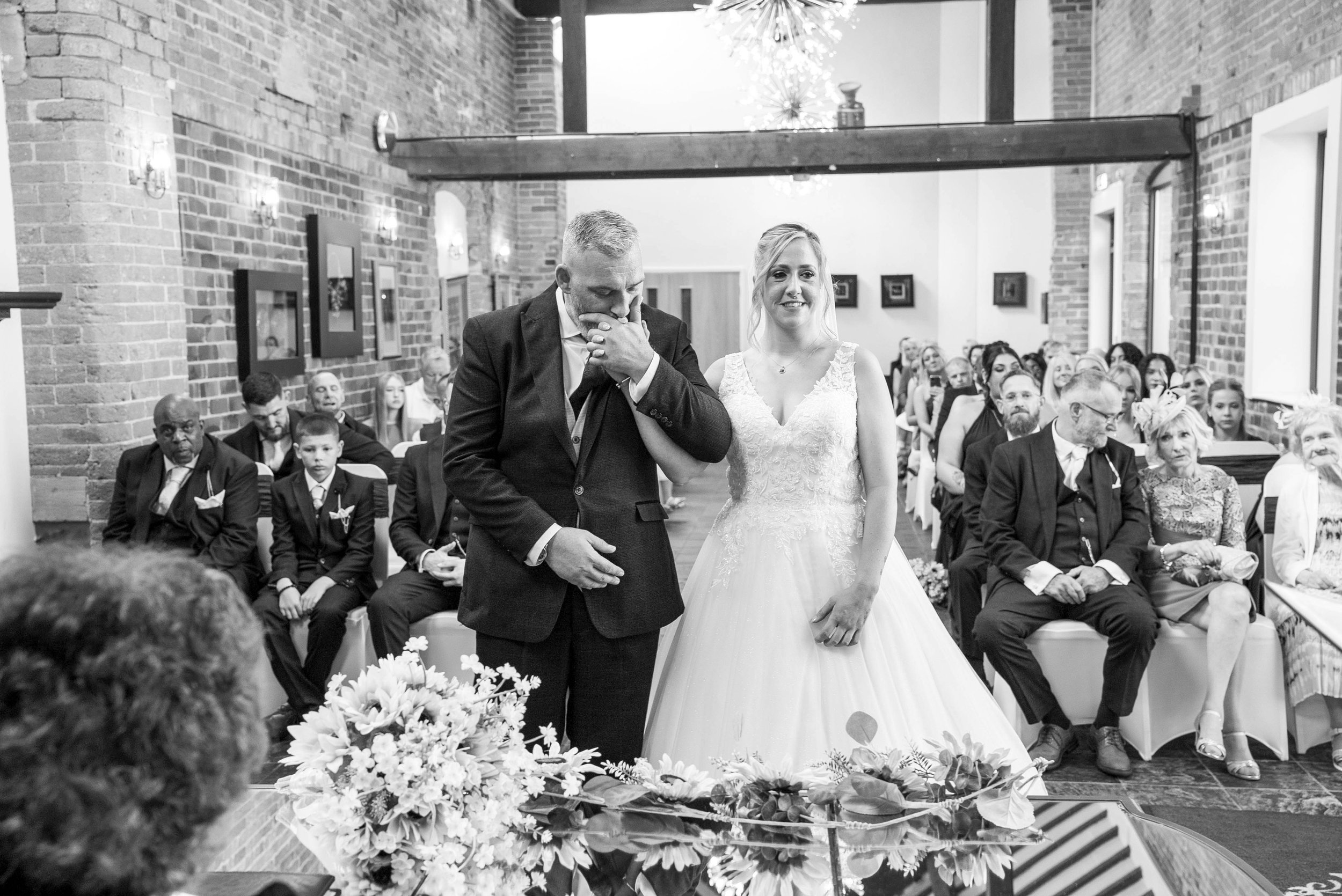A bride and her father at a wedding ceremony, with the father wiping tears from his eyes and the bride smiling, in a room with exposed brick walls and seated guests in the background.